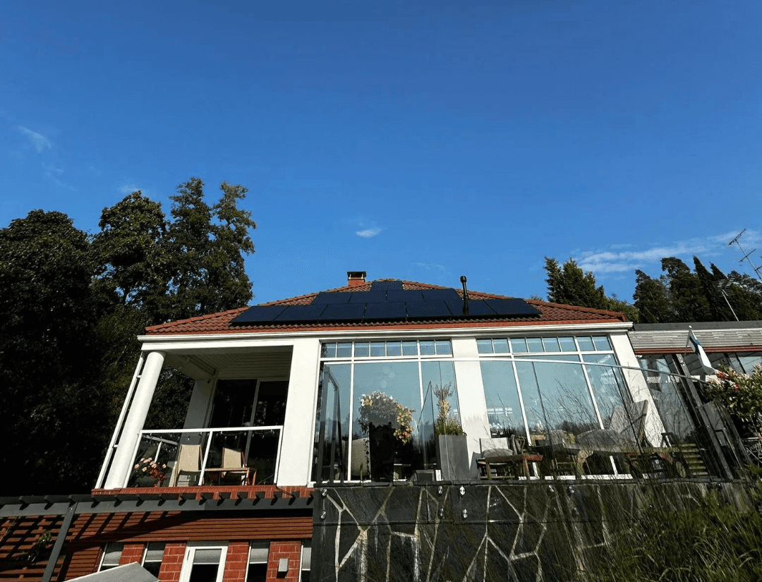 Talo, jossa on lasitettu veranta ja patio, punatiilikatolla aurinkopaneelit, taustalla sininen taivas ja puita.