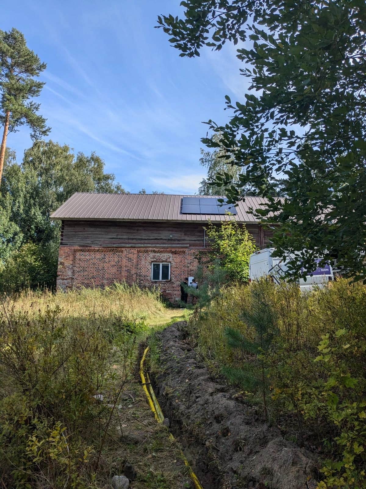 Tiilestä ja puusta rakennettu rakennus maaseutumaisessa, rehevässä ympäristössä, jonka katolle on asennettu aurinkopaneelit. Rakennusta kohti kulkee kaivettu kaivanto, jossa näkyy kaapeli, ympärillä puita ja kasvillisuutta.