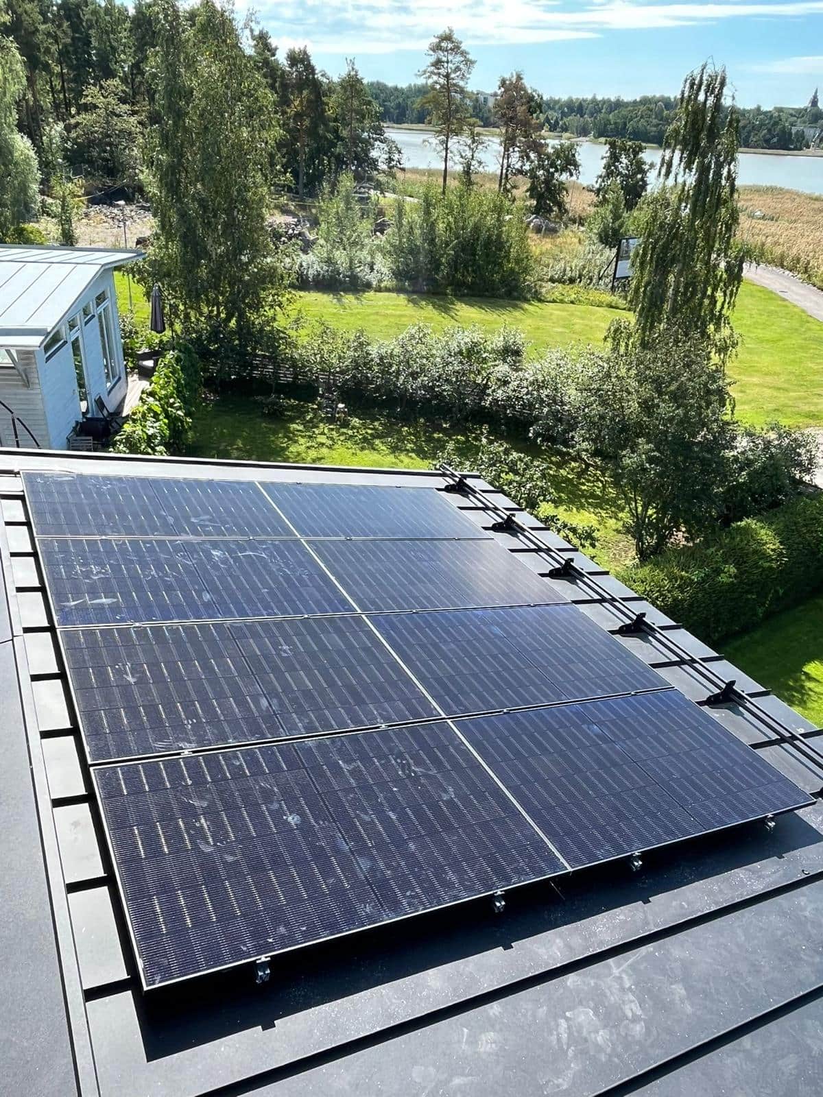 Lähikuva omakotitalon viistokatolle asennetuista aurinkopaneeleista, joissa näkyvät kiinnityskiskot sekä taustalla vihreä piha ja puita.