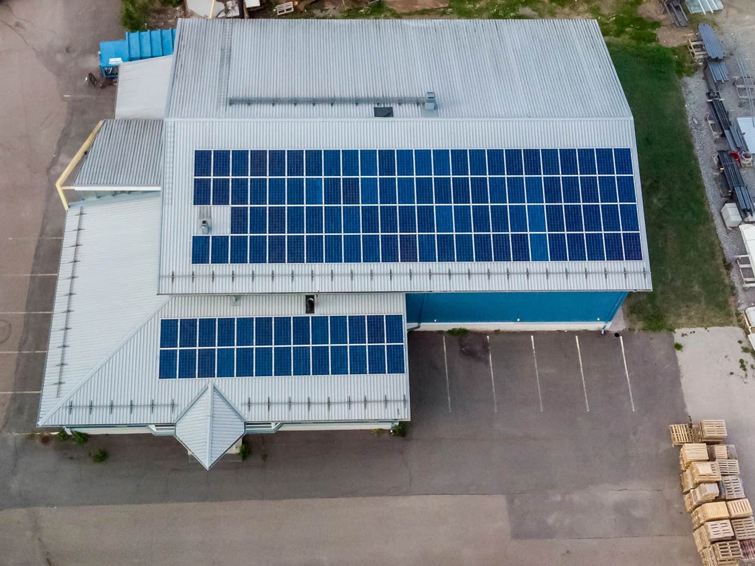 Ilmakuva metallikattoisesta liikerakennuksesta, jonka katto on peitetty suorakulmaisilla aurinkopaneeleilla, vieressä pysäköintialue ja pinottuja lavoja.