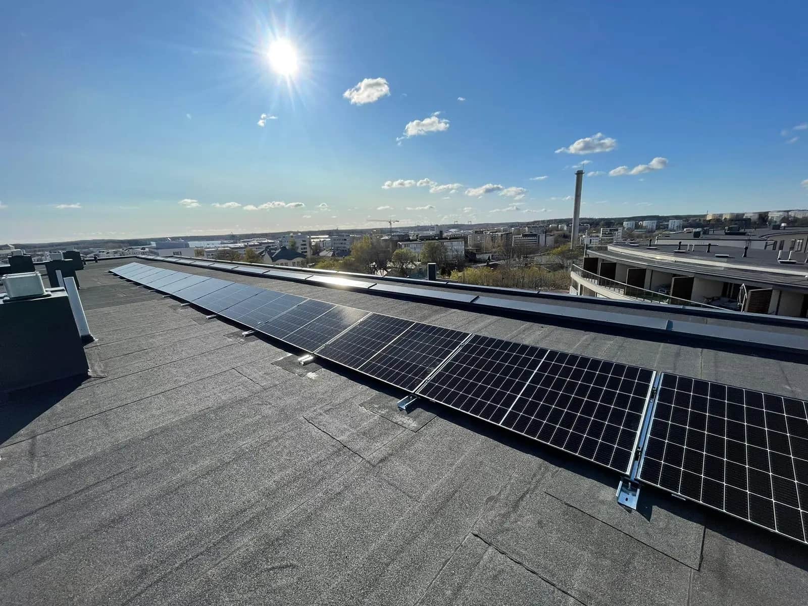 Aurinkopaneelit tasakatolla kirkkaan sinisen taivaan ja kirkkaan auringon alla, taustalla siintää kaupunkimaisema.