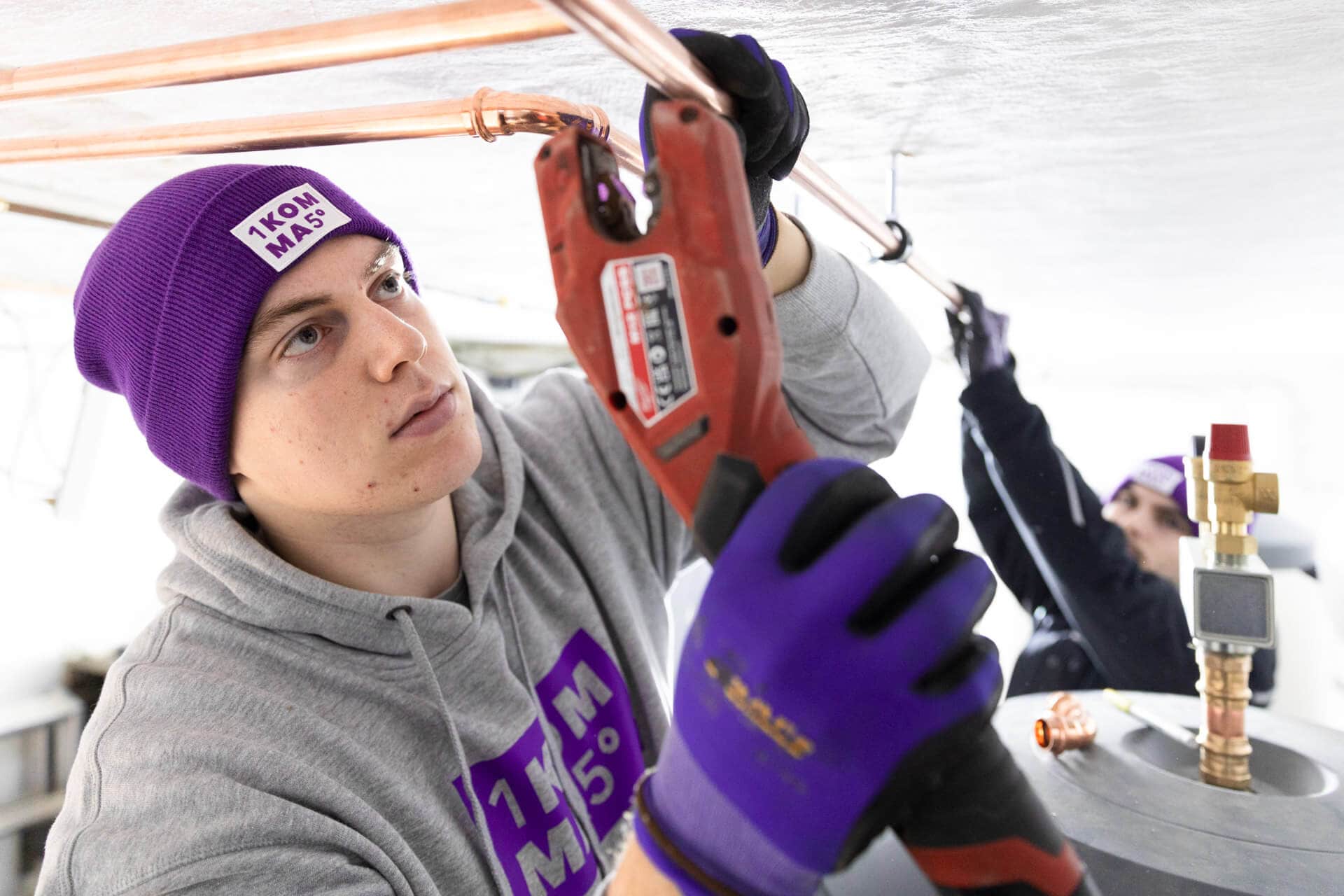 Asentaja suojakäsineissä ja pipossa asentaa kupariputkia sähkötyökalulla sisätiloissa, toisen asentajan työskennellessä taustalla.