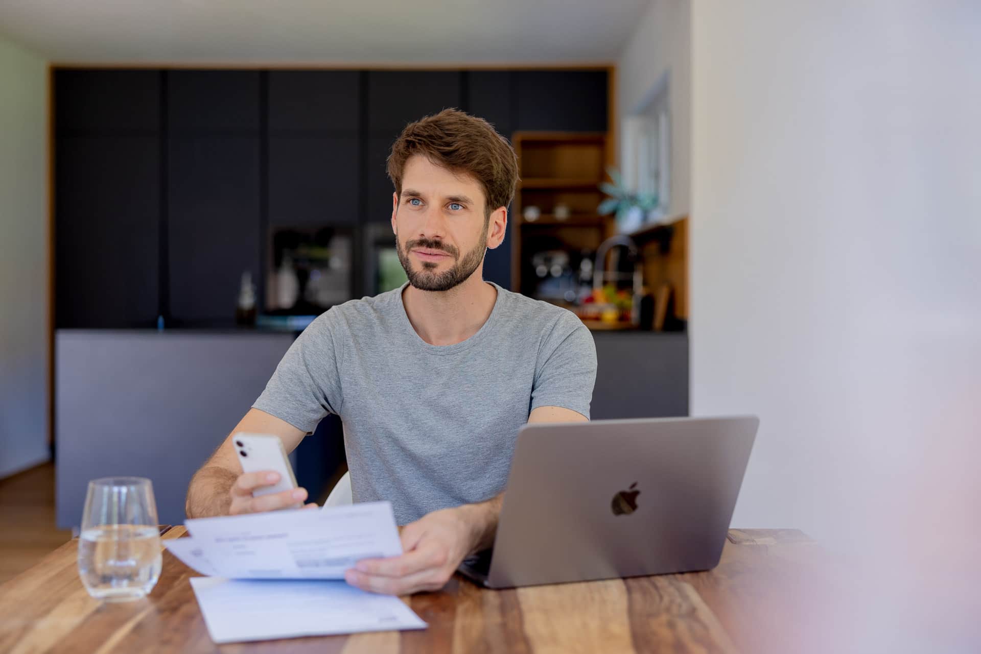 Mies istuu pöydän ääressä kannettavan tietokoneen kanssa, pitäen kädessään älypuhelinta ja asiakirjoja, modernissa keittiöympäristössä.