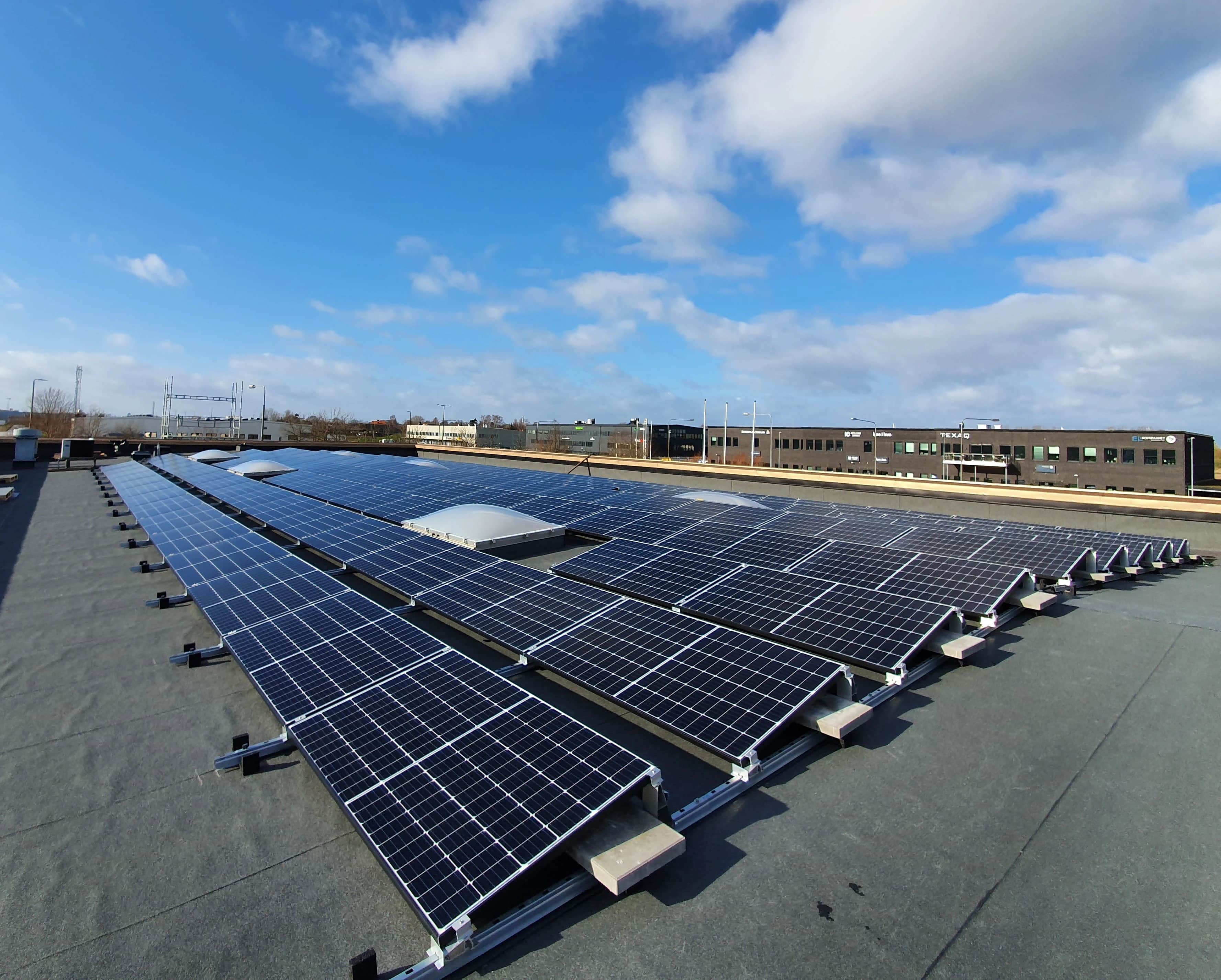 Riveittäin aurinkopaneeleita tasaisella liikerakennuksen katolla kirkkaan sinisen, pilviharson peittämän taivaan alla.