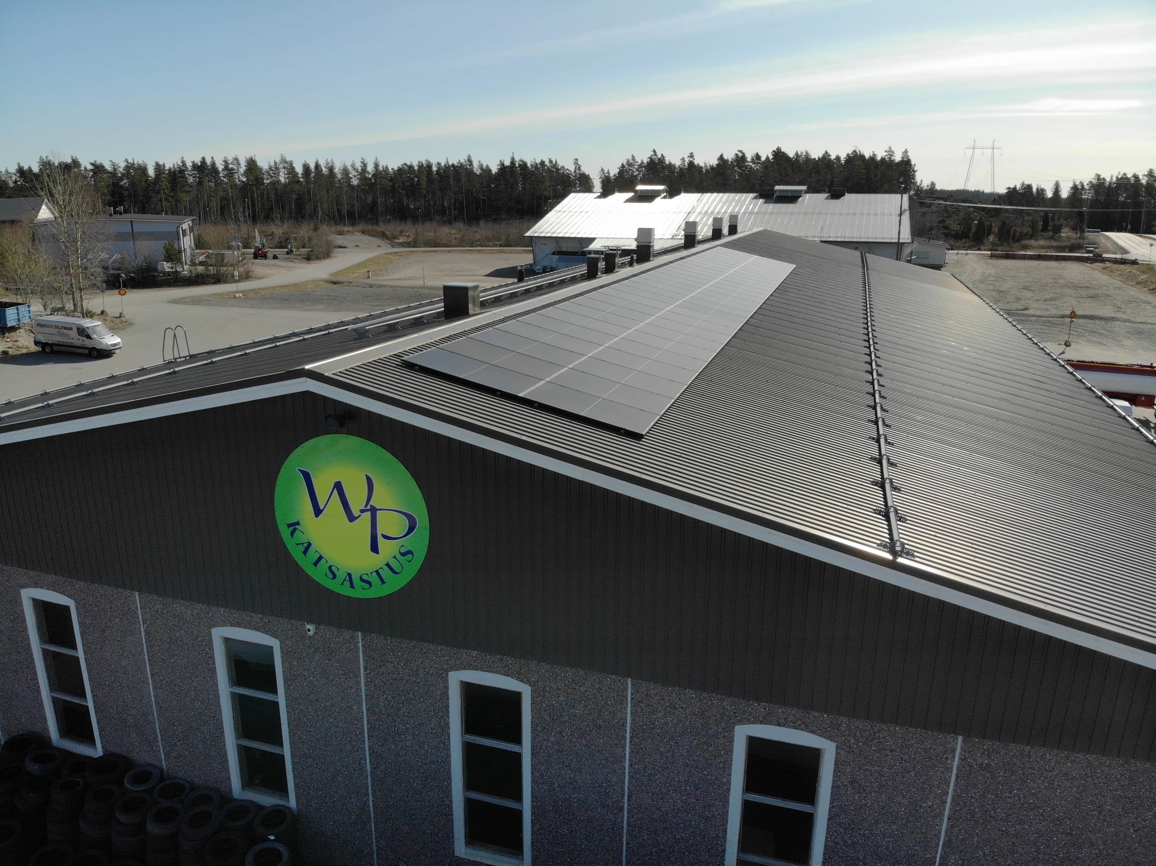 Metallikattoinen teollisuusrakennus, jonka katolla on aurinkopaneelit ja etuosassa vihreä pyöreä "WP"-logo, taustalla puita.