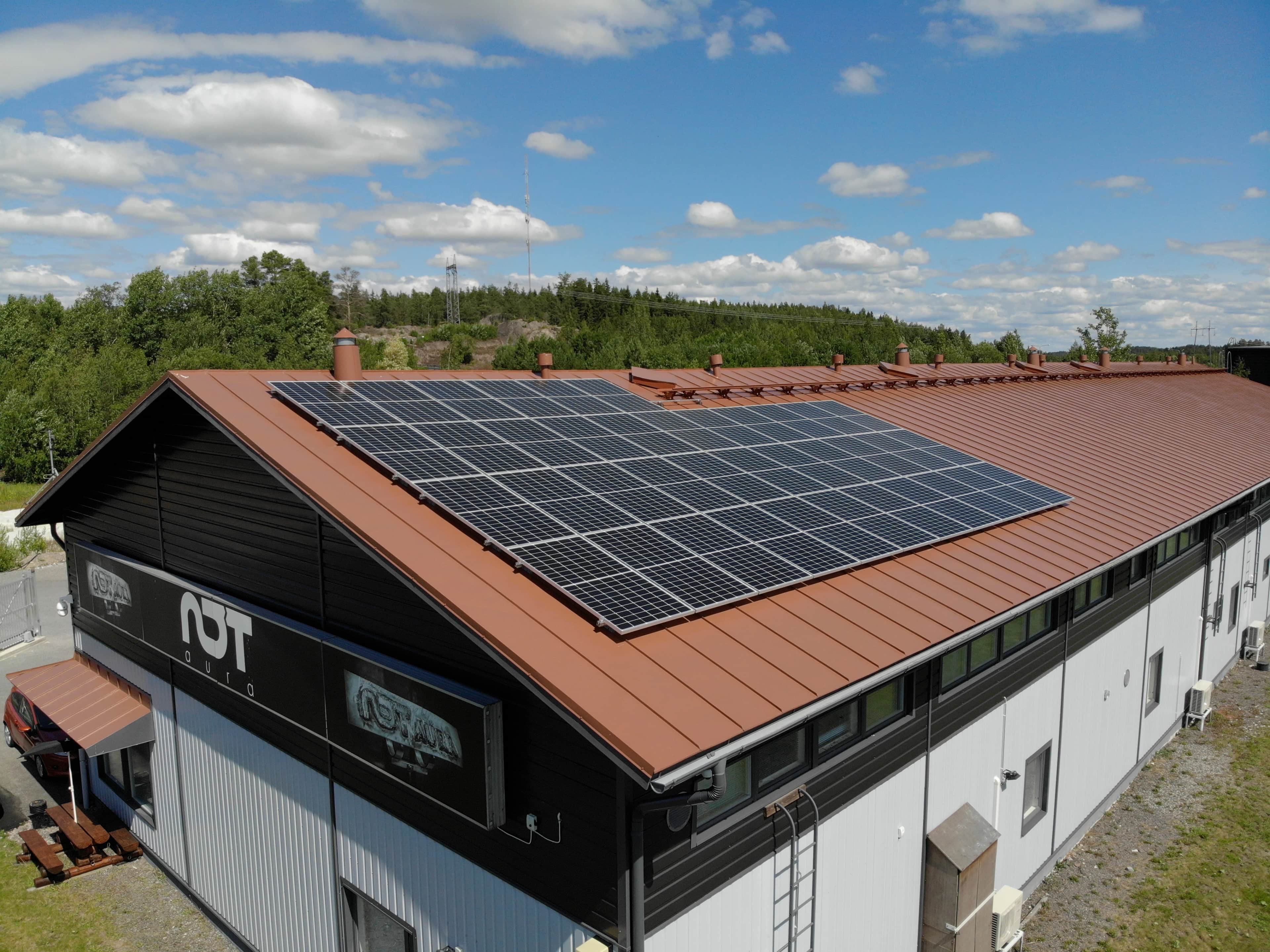 Teollisuusrakennus, jonka punainen metallikatto on peitetty aurinkopaneeleilla, sijaiten puiden keskellä sinisen, pilviharsoisen taivaan alla.