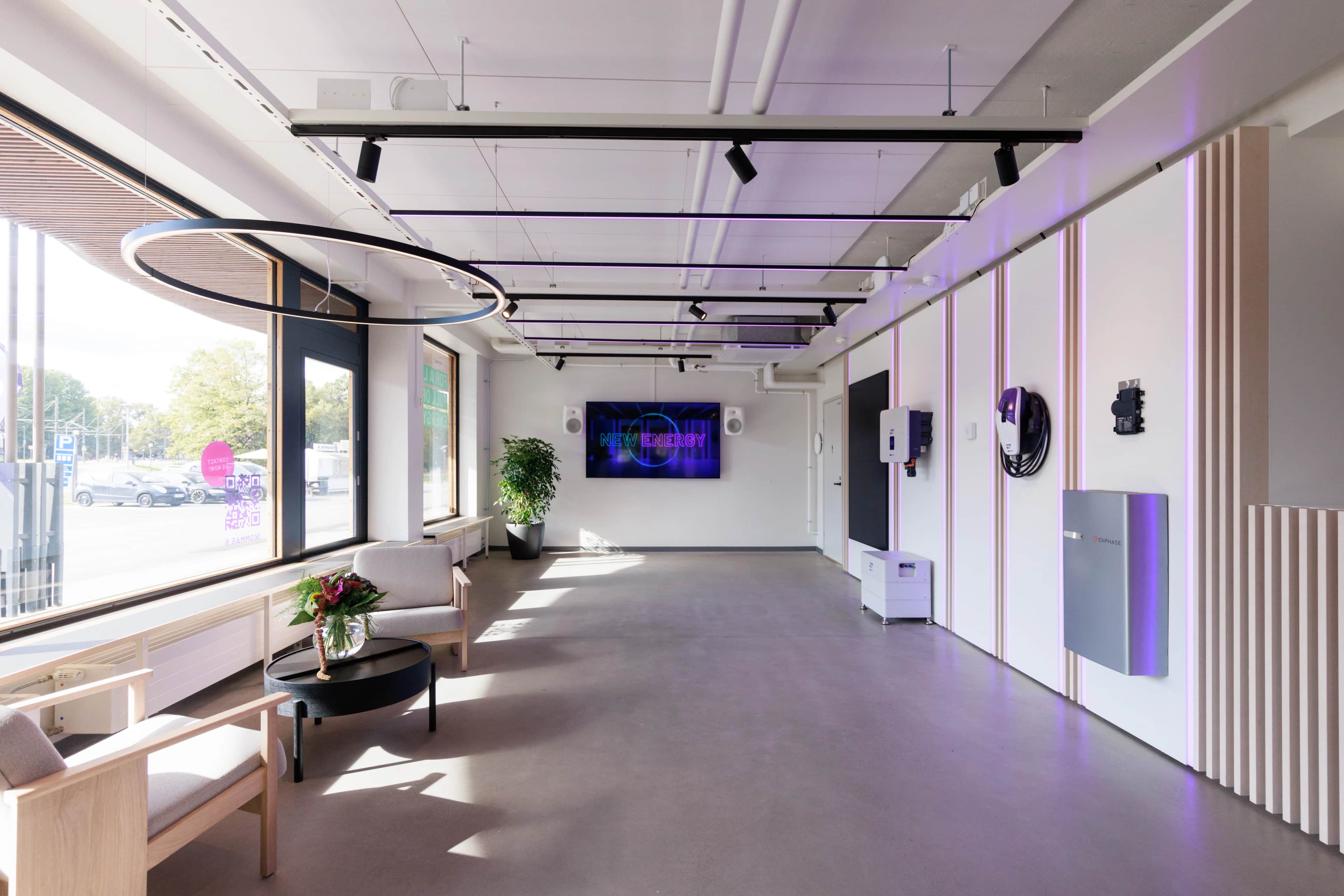 Valoisa moderni esittelytila, jossa on suuret ikkunat, pyöreä riippuvalaisin, oleskelualue, viherkasvi ruukussa, seinälle asennettu näyttö ja violetit korostusvalot.