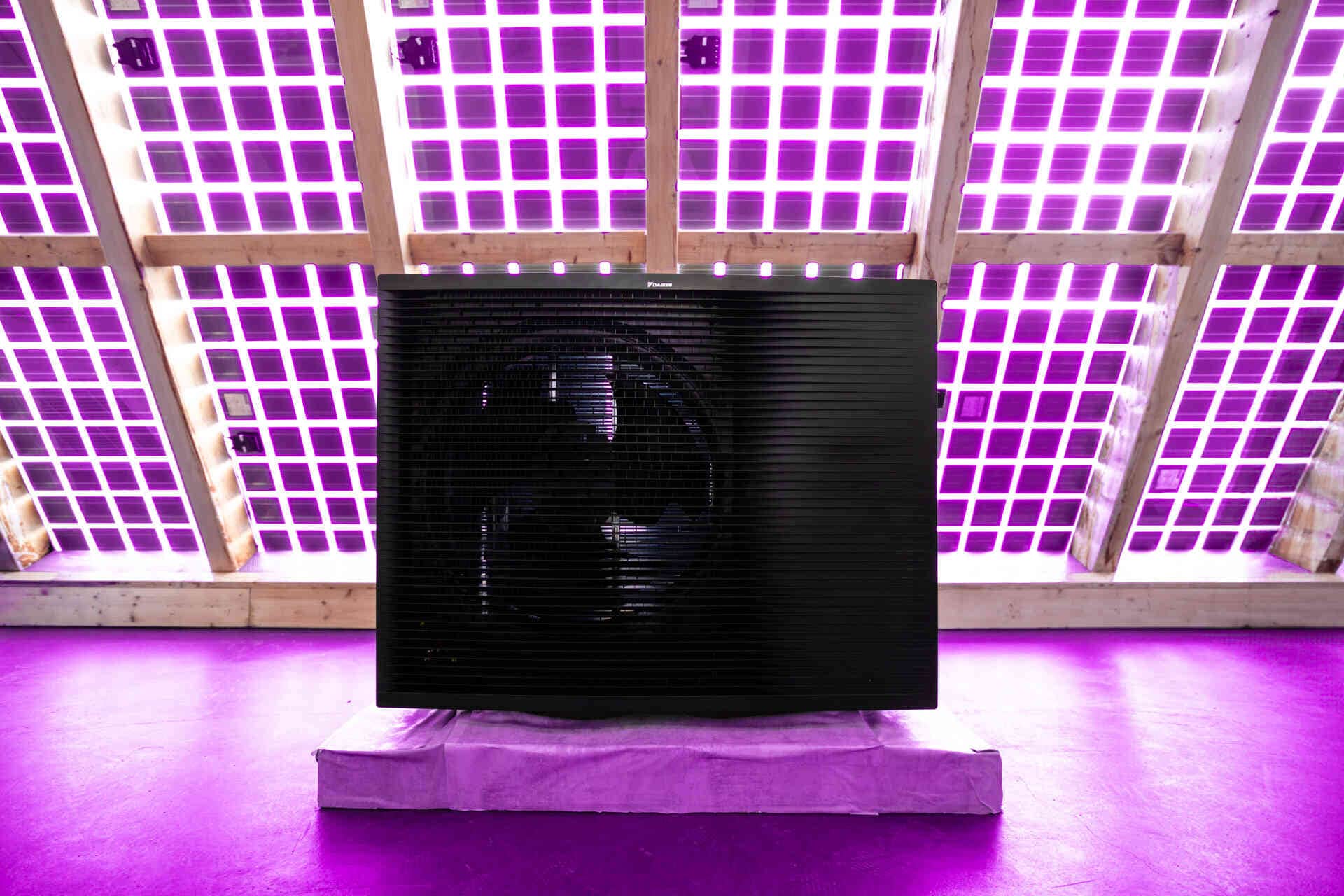 Musta lämpöpumppuyksikkö esillä sisätiloissa, taustalla violetit LED-valot ja ritilämäinen katto.