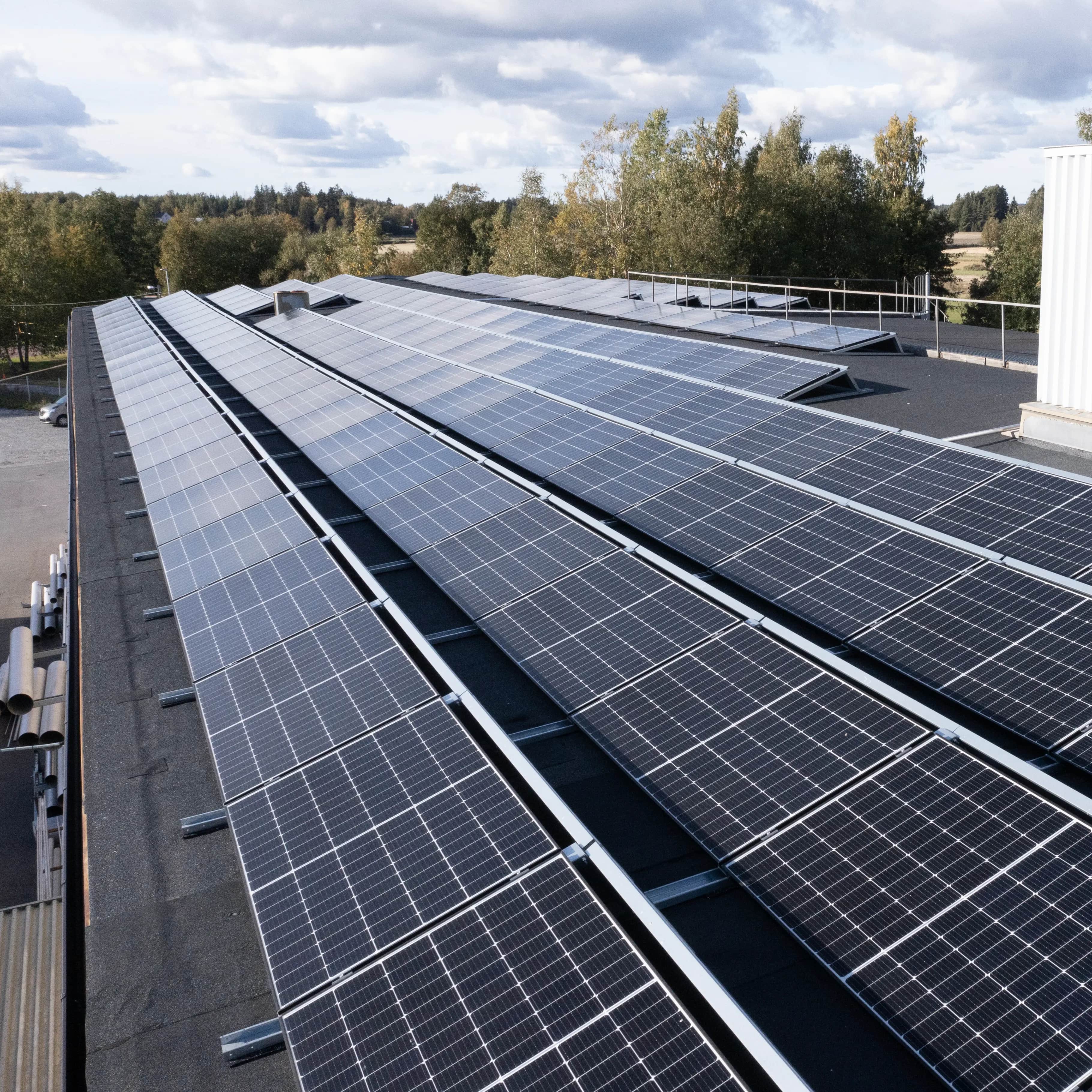 Useita rivejä aurinkopaneeleita tasakatolla, taustalla puita ja pilvinen taivas.
