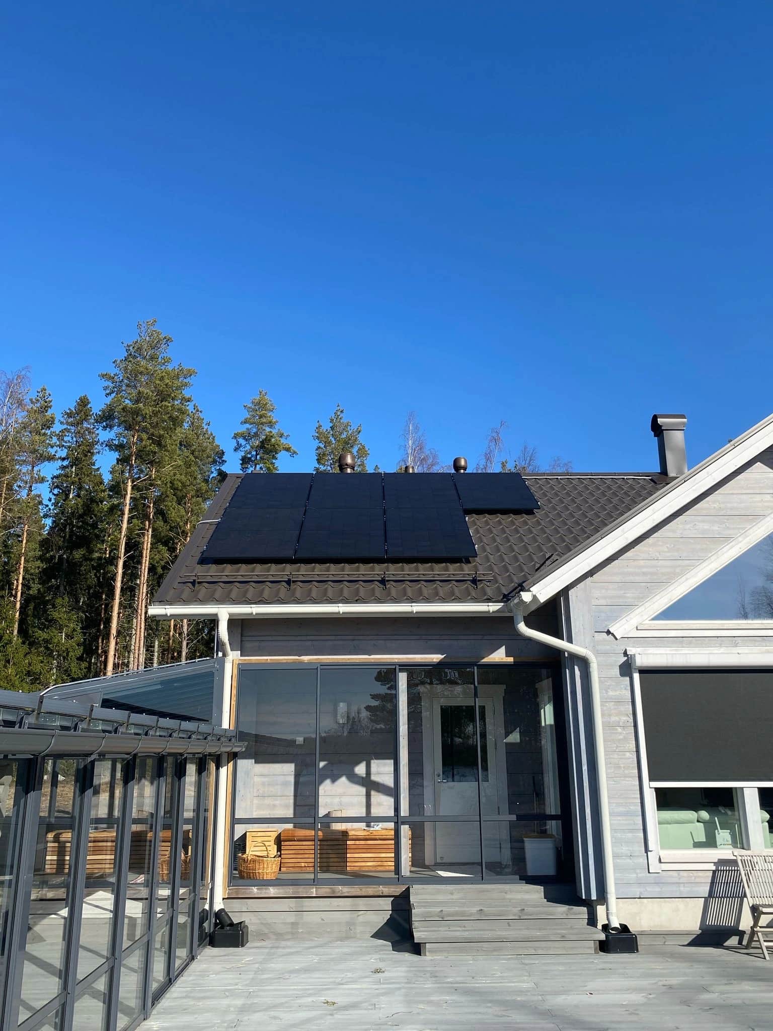 Moderni talo, jonka katolla on aurinkopaneelit, puiden ympäröimänä kirkkaan sinisen taivaan alla. Etualalla näkyy lasitettu terassi.