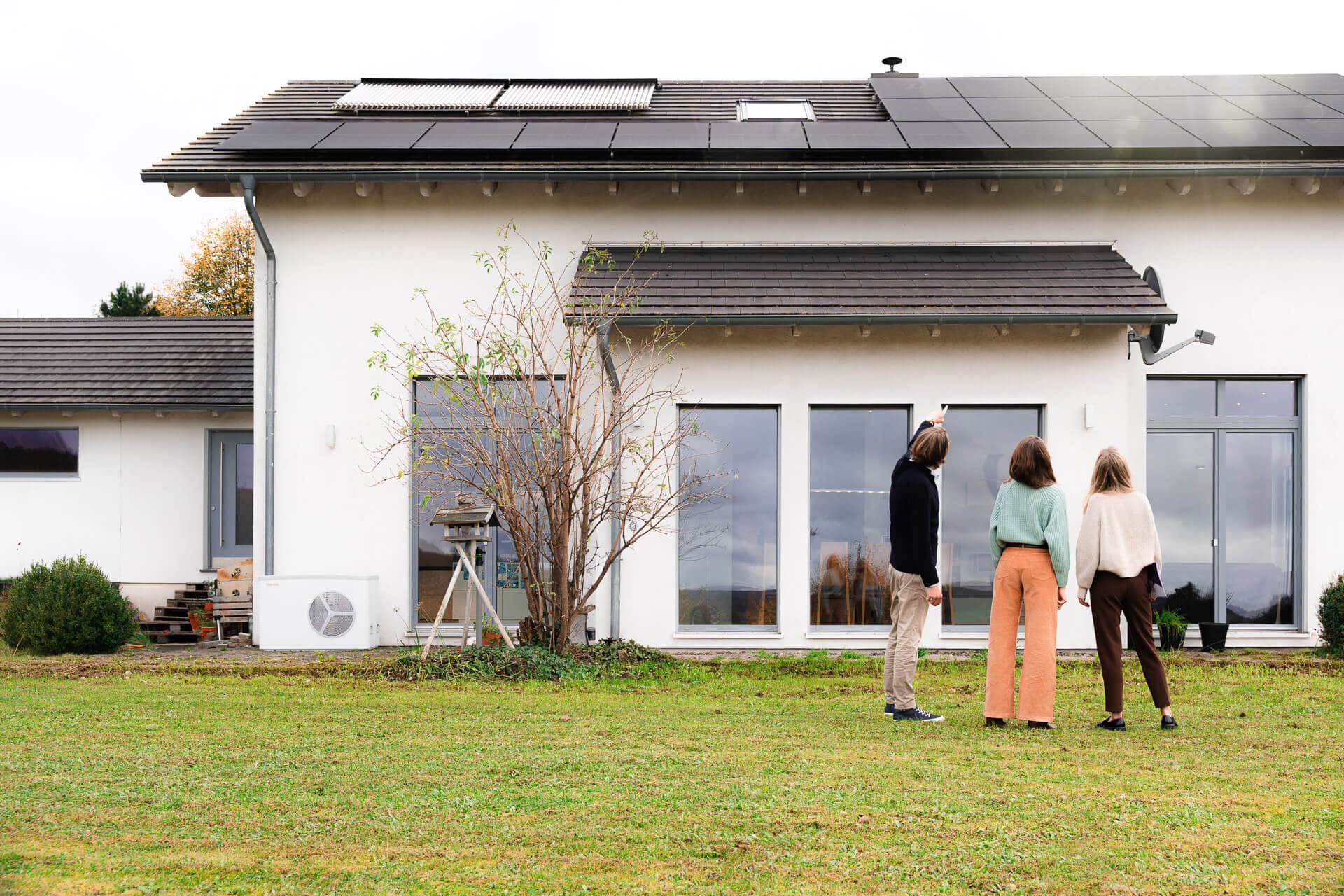 Kolme ihmistä seisoo modernin valkoisen talon edessä, jonka katolla on aurinkopaneelit, katsellen rakennusta.