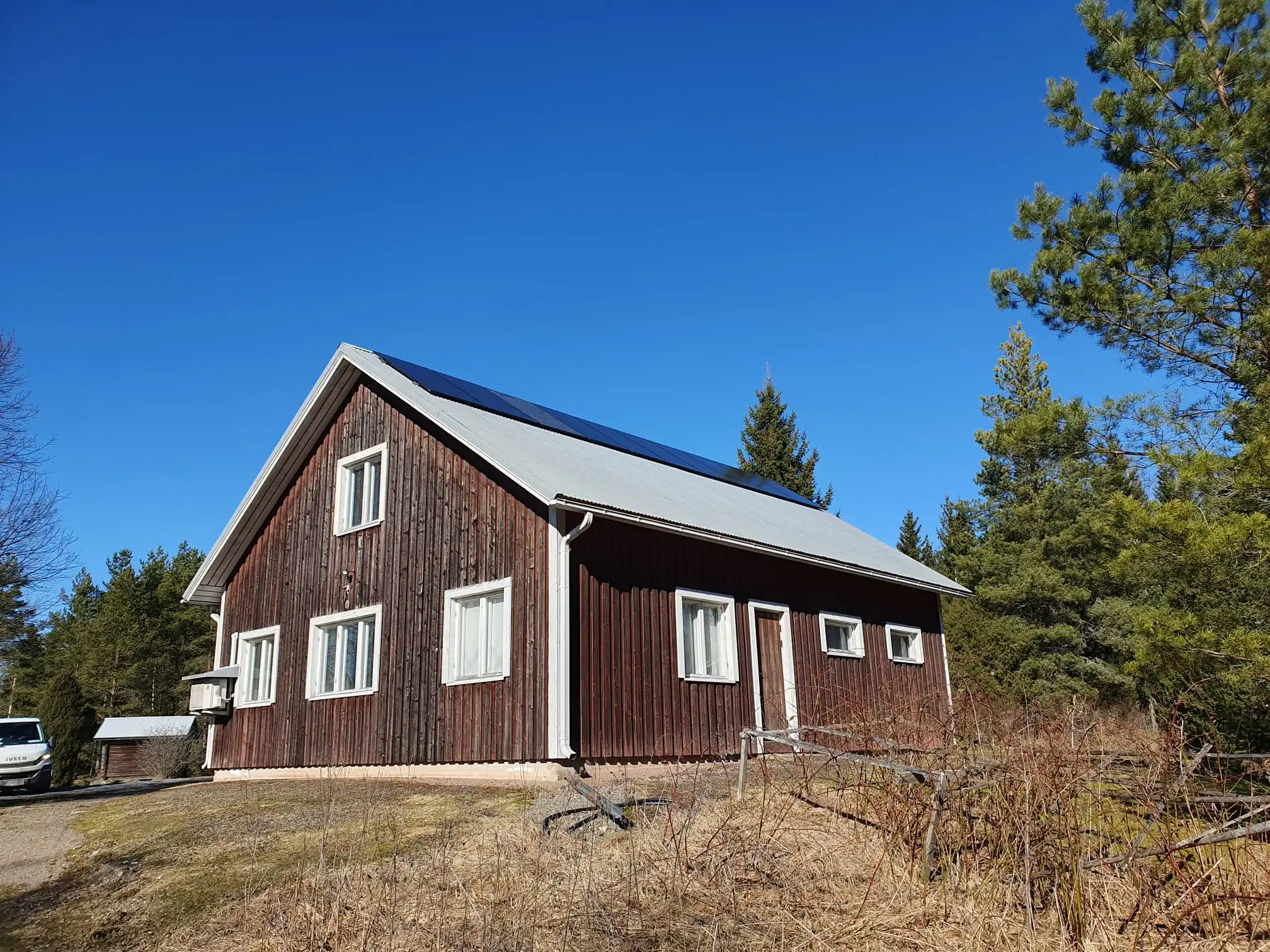 Ulkokuva puisesta omakotitalosta maaseutumaisessa ympäristössä, jossa katolle on asennettu aurinkopaneelit ja taloa ympäröi metsää kirkkaan sinisen taivaan alla.