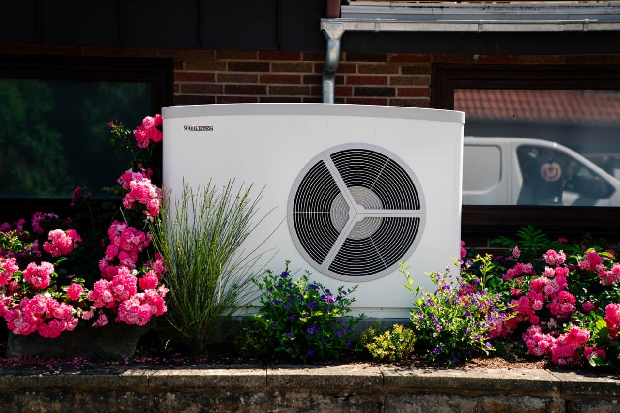 Valkoinen lämpöpumppuyksikkö on värikkäiden pinkkien kukkien ja vehreyden ympäröimä, taustalla tiiliseinä, jossa on ikkuna, ja pakettiauto.