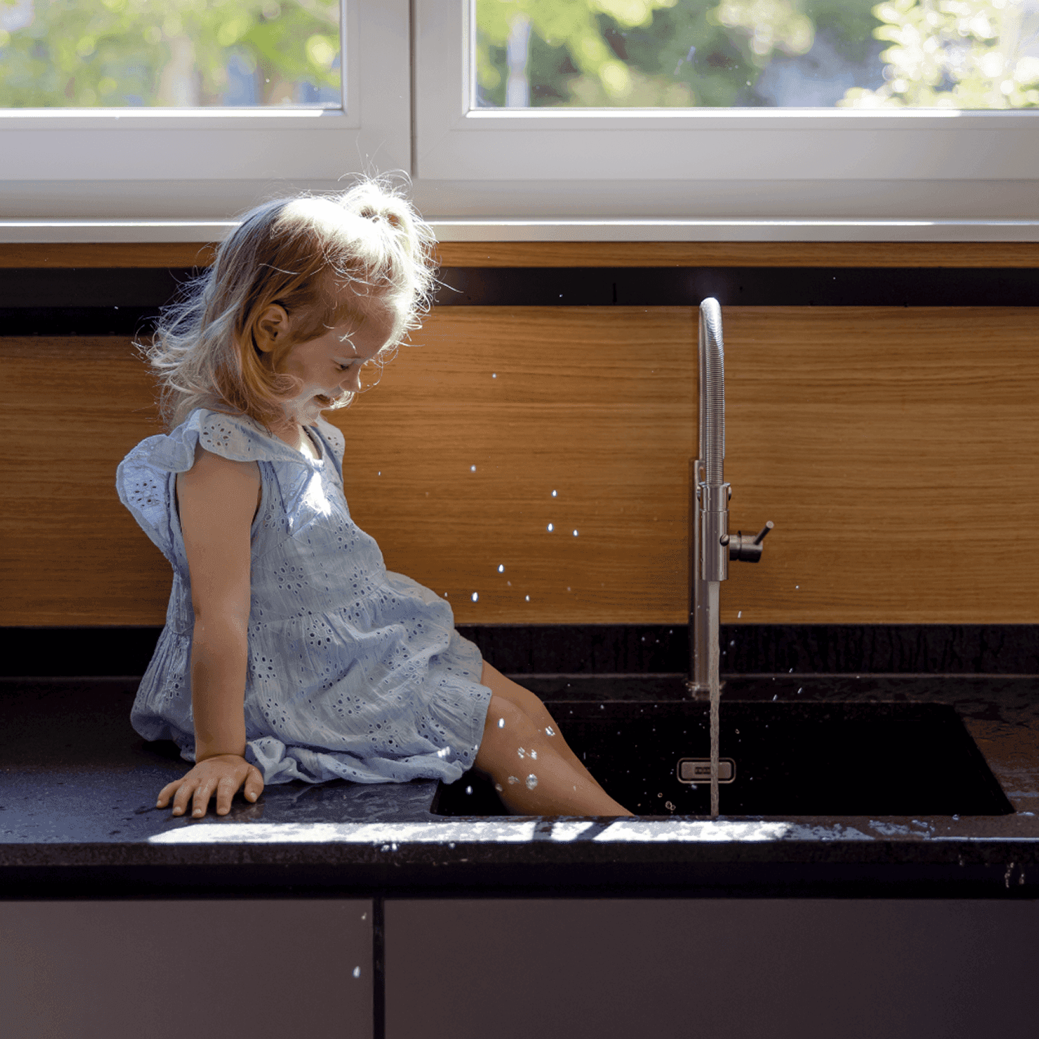 Hymyilevä nainen tummansinisessä neuleessa istuu ikkunan ääressä; taustalla kolme ihmistä keskustelee pöydän ääressä.