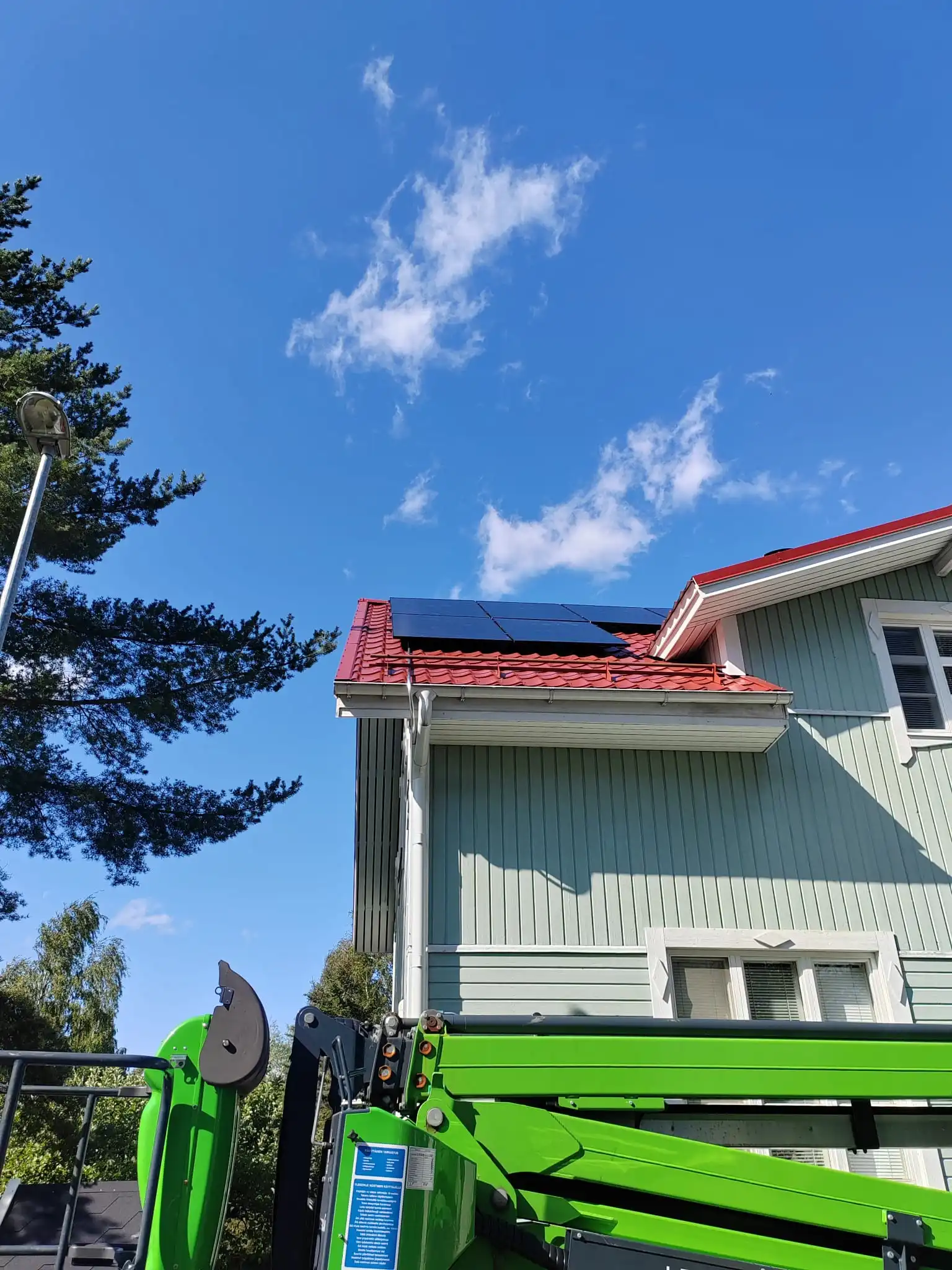 Vaaleanvihreä talo, jossa on punainen katto ja aurinkopaneelit; etualalla vihreä nostin, korkea mänty ja lyhtypylväs kirkkaan sinisen, pilviharsoisen taivaan alla.