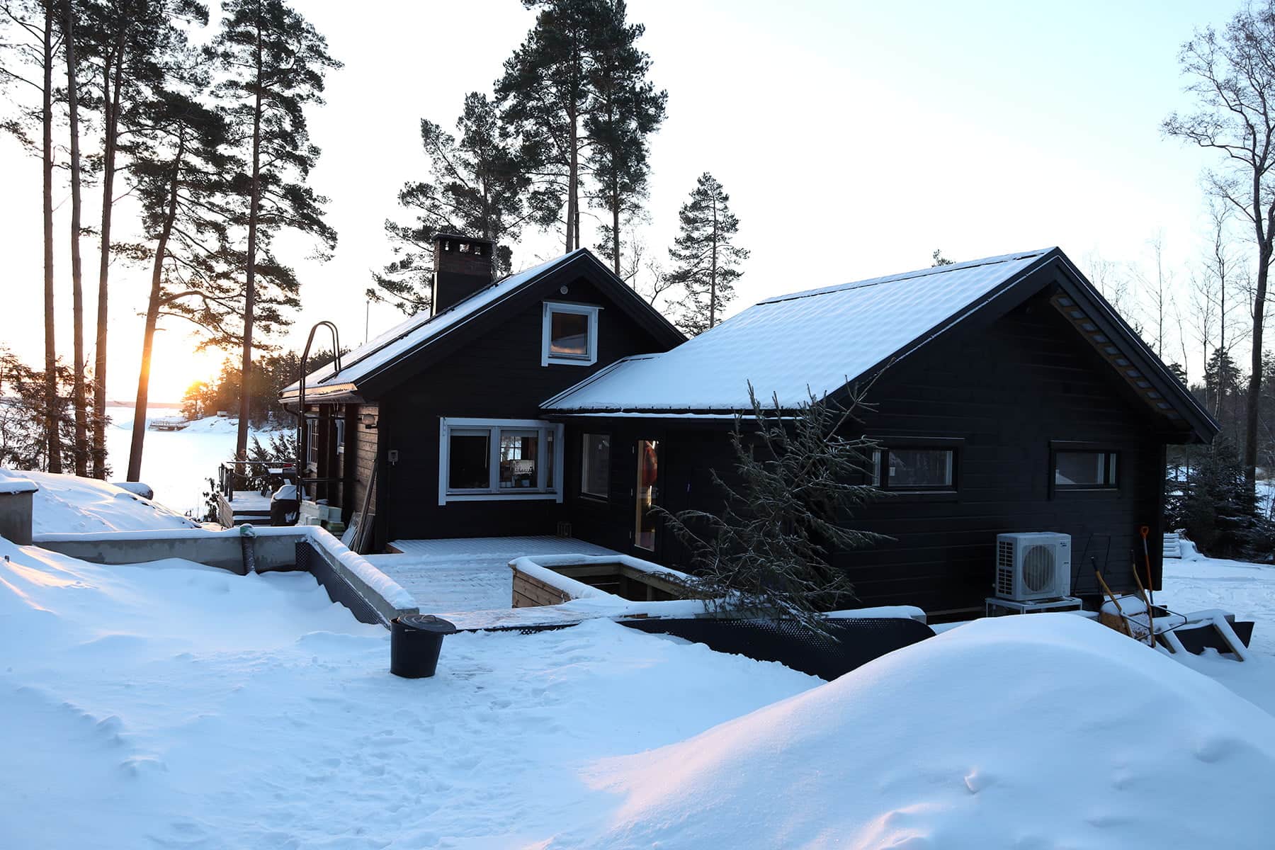 Tumma hirsimökki lumen ympäröimänä, taustalla korkeita mäntyjä ja järvi kirkkaan taivaan alla, auringon laskiessa horisonttiin.