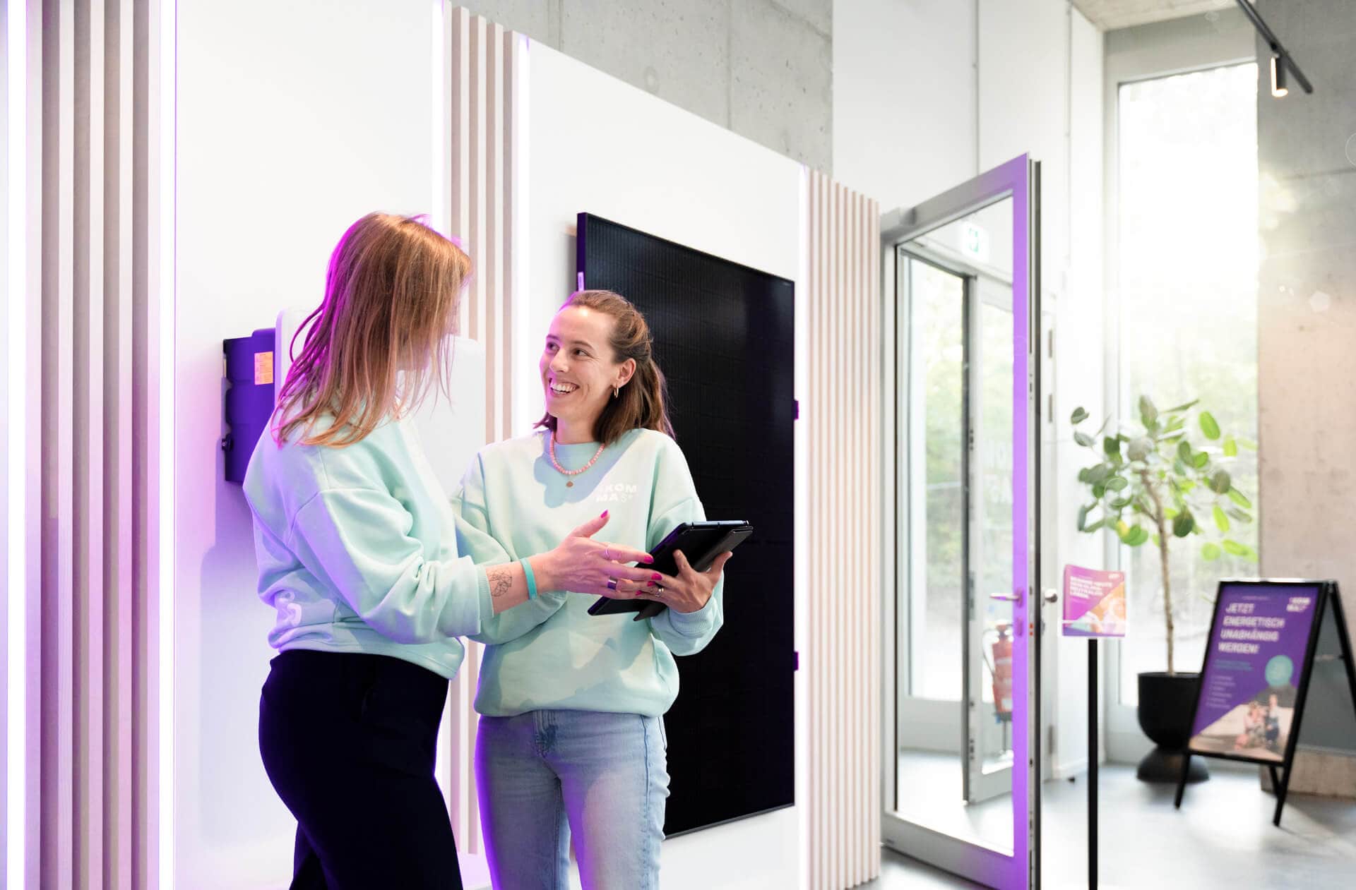 Kaksi vaaleisiin collegepaitoihin pukeutunutta naista hymyilee ja keskustelee, toisen pidellessä tablettia modernissa, violetisti valaistussa toimiston aulassa.