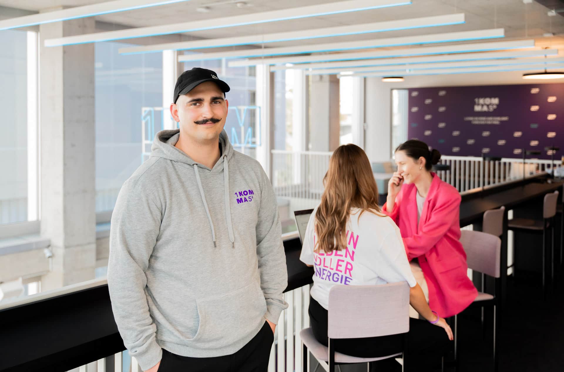 Viiksellinen ja lippalakkinen mies harmaassa hupparissa seisoo valoisassa modernissa toimistossa, taustalla kaksi naista juttelee korkean pöydän äärellä.