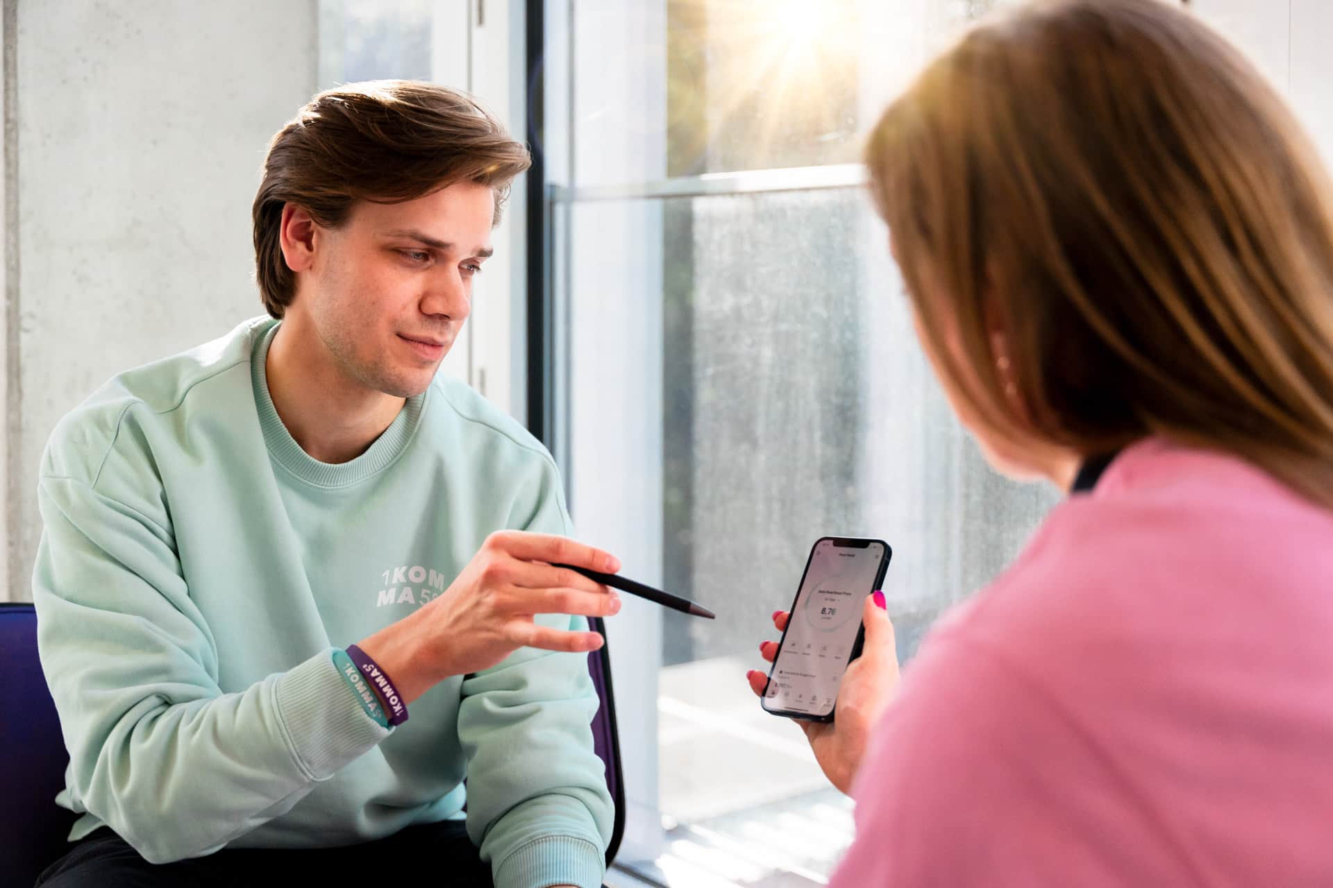 Vihreään collegepaitaan pukeutunut mies osoittaa kynällä älypuhelimen näyttöä, jota vaaleanpunaiseen pukeutunut nainen pitelee, heidän istuessaan auringonvalossa ikkunan äärellä.