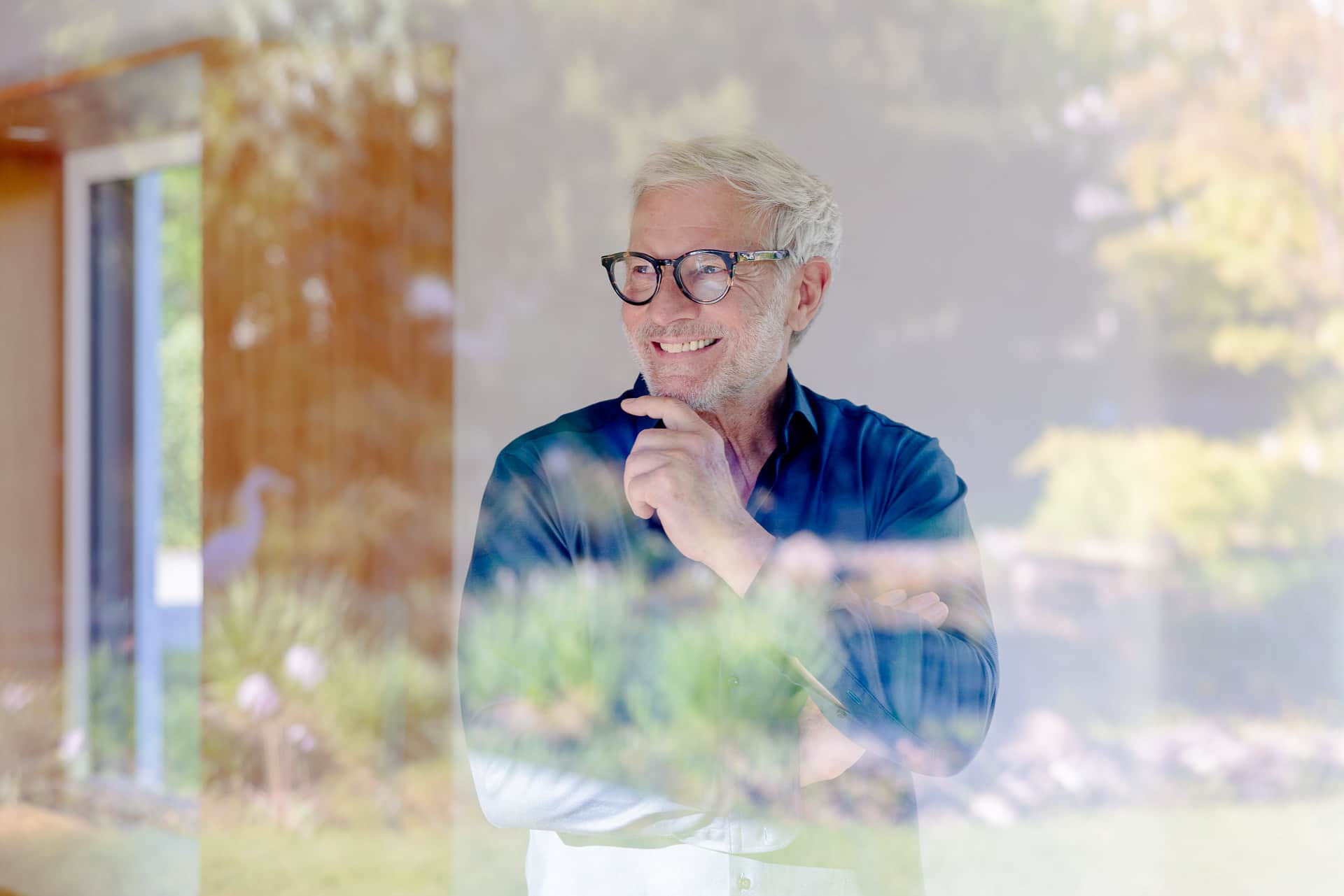 Silmälasipäinen mies hymyilee seisten sisätiloissa; ikkunan pinnasta heijastuvat puut ja puutarha luovat kerroksellisen vaikutelman.