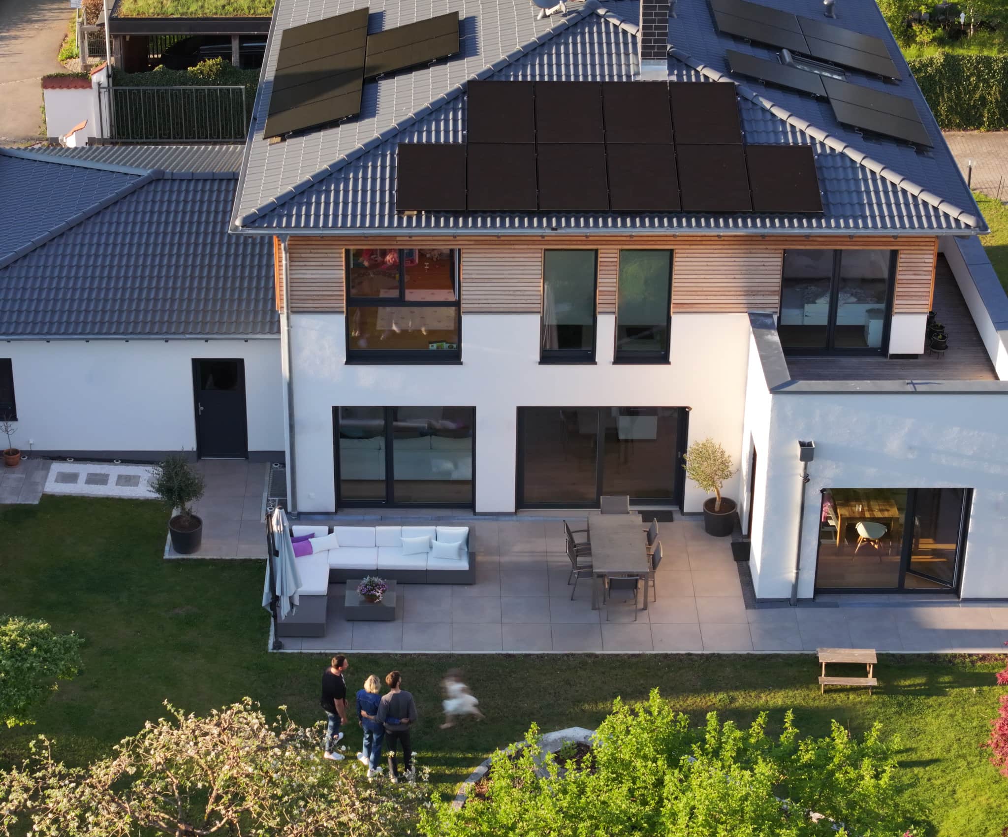Ilmakuva modernista talosta, jonka katolla on aurinkopaneelit, patiolla ulkokalusteita ja puutarhassa ihmisiä kokoontuneina.