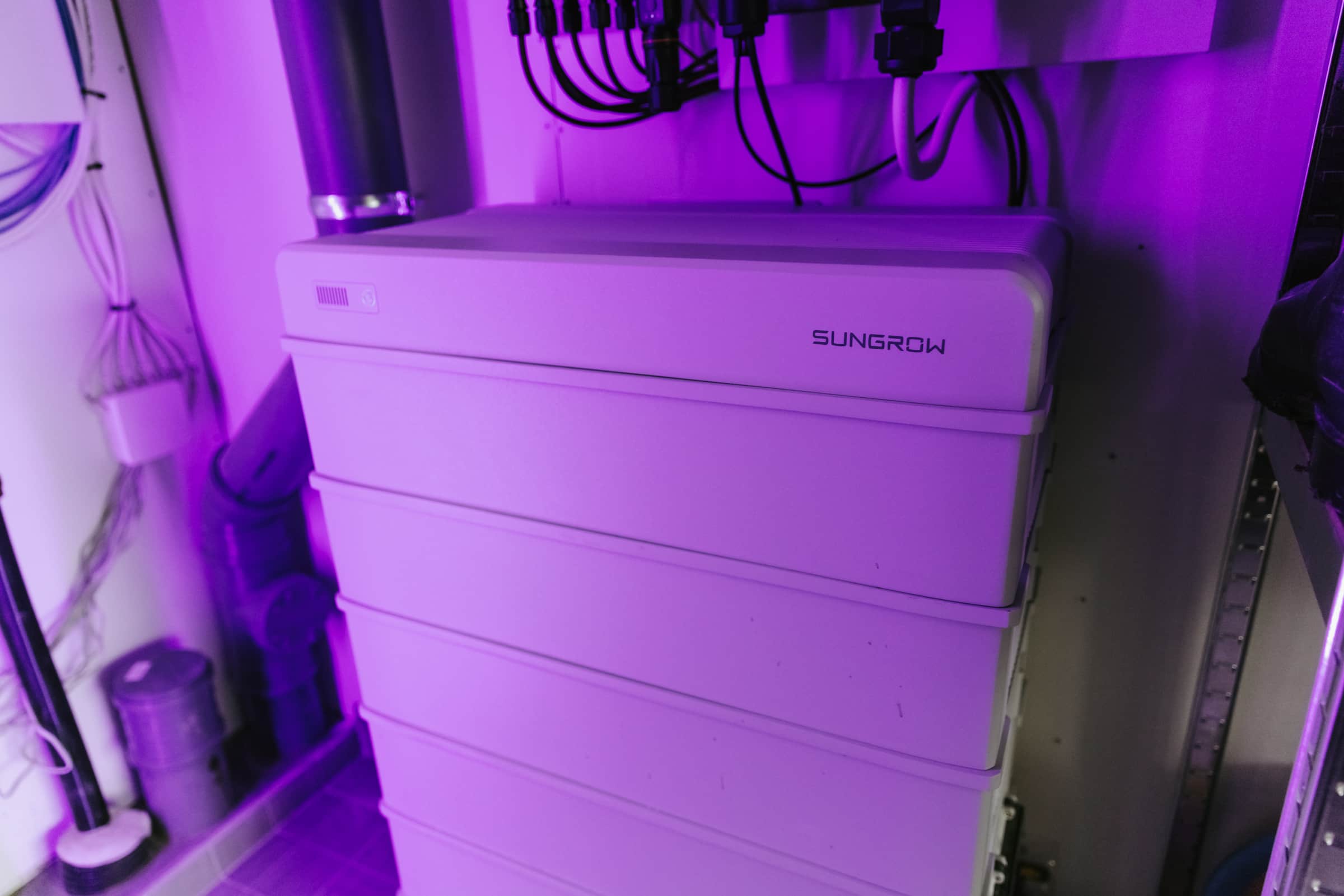 Valkoinen Sungrow-akkujärjestelmä huoneessa, jossa on violetti valaistus, ympärillä kaapeleita ja laitteita.