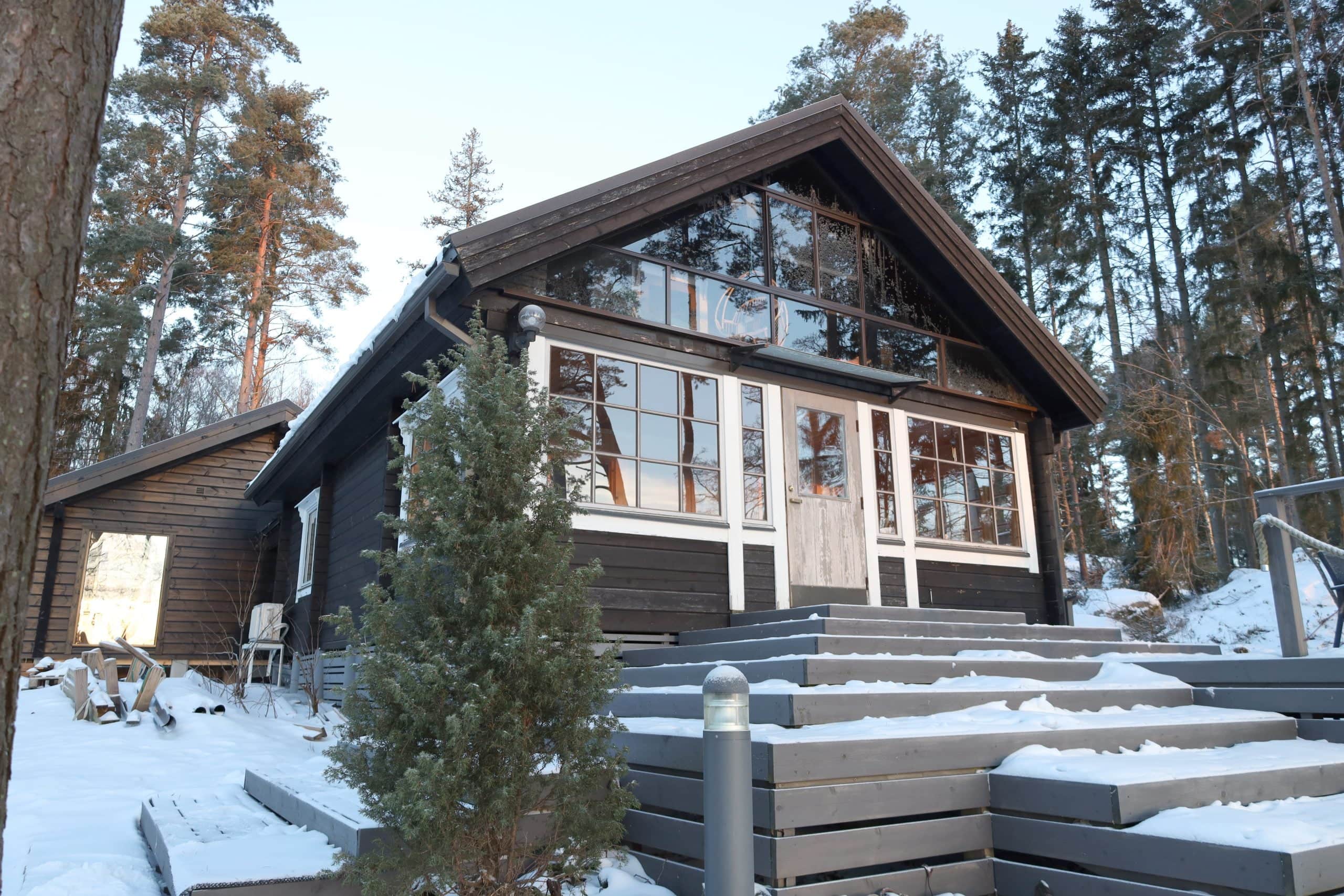 Hirsimökki, jossa on suuret ikkunat, lumisen maiseman ja puiden ympäröimänä, varustettuna portaittaisella sisäänkäynnillä ja etualalla pienellä havupuulla. Oikealla puolella on asennettu lämpöpumppu.