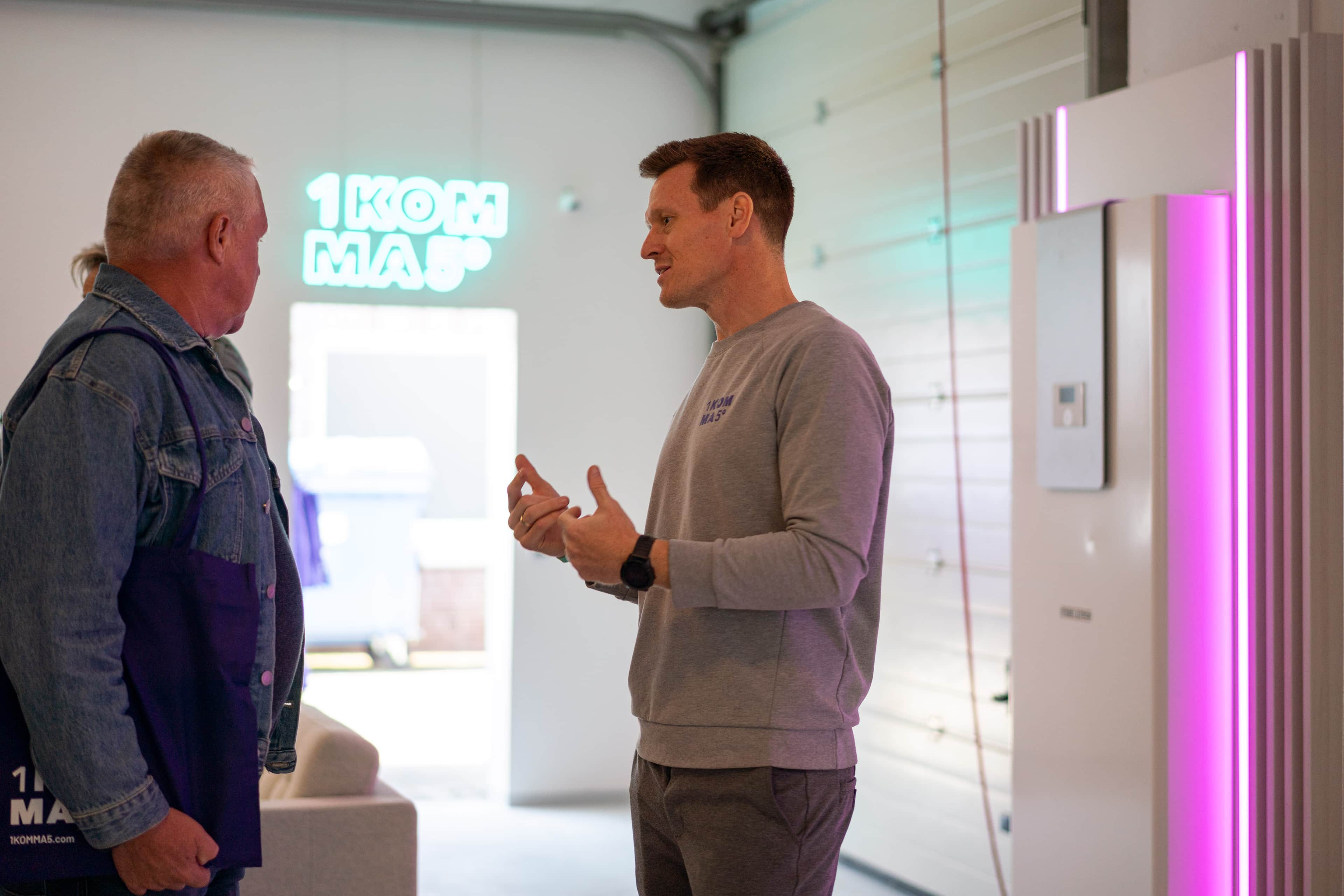 Kaksi miestä keskustelevat modernissa huoneessa, jossa on neonkylttejä; toisella on farkkutakki ja toisella harmaa neule. Lähettyvillä hohtaa vaaleanpunainen valo.