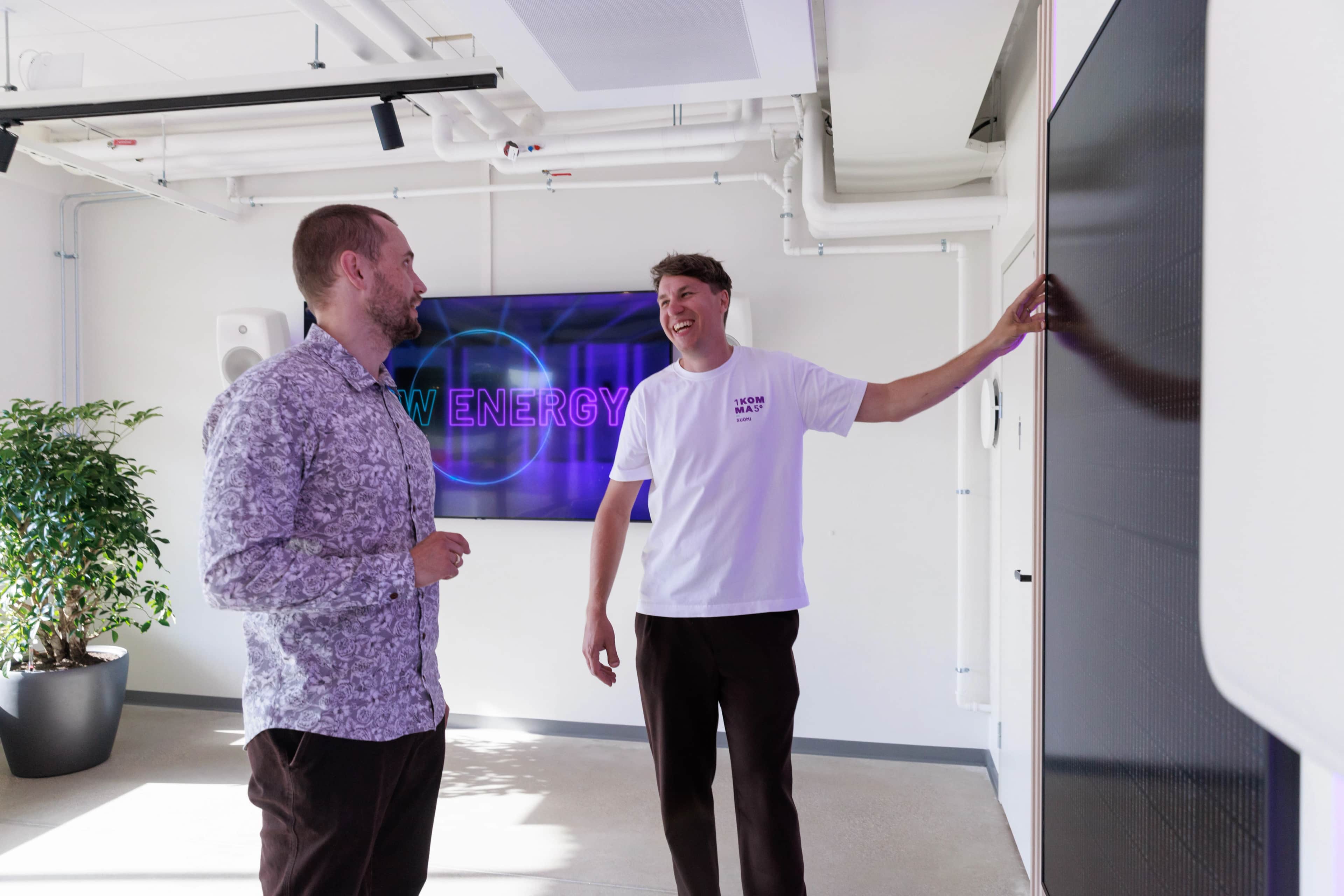 Kaksi miestä modernissa toimistotilassa keskustelevat suuren näytön äärellä; toinen osoittaa näyttöä, ja taustalla näkyy kasvi sekä “New Energy” -kyltti.