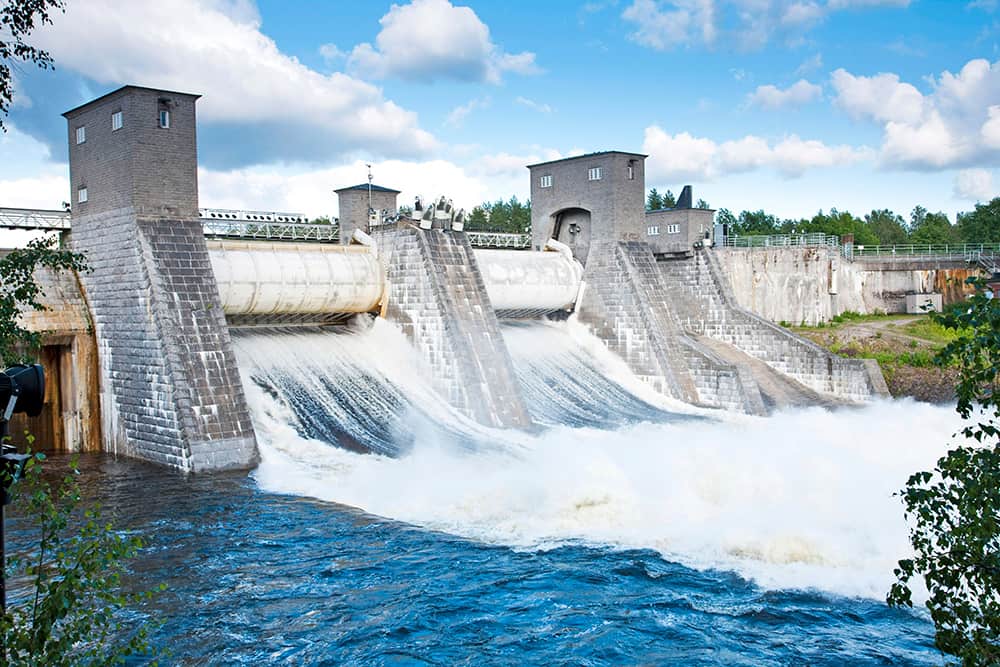 Suuri vesivoimalaitos, jossa on useita sulkuportteja vapauttamassa vettä jokeen, puiden ympäröimänä osittain pilvisen taivaan alla.