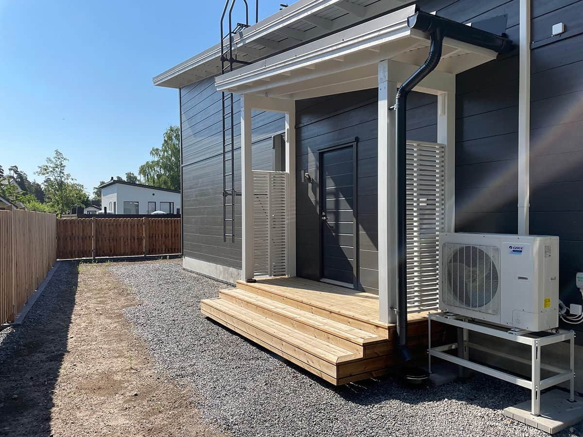 Moderni talon julkisivu, jossa on pieni puinen kuisti, lämpöpumppuyksikkö ja sorapolku, kirkkaan sinisen taivaan alla.