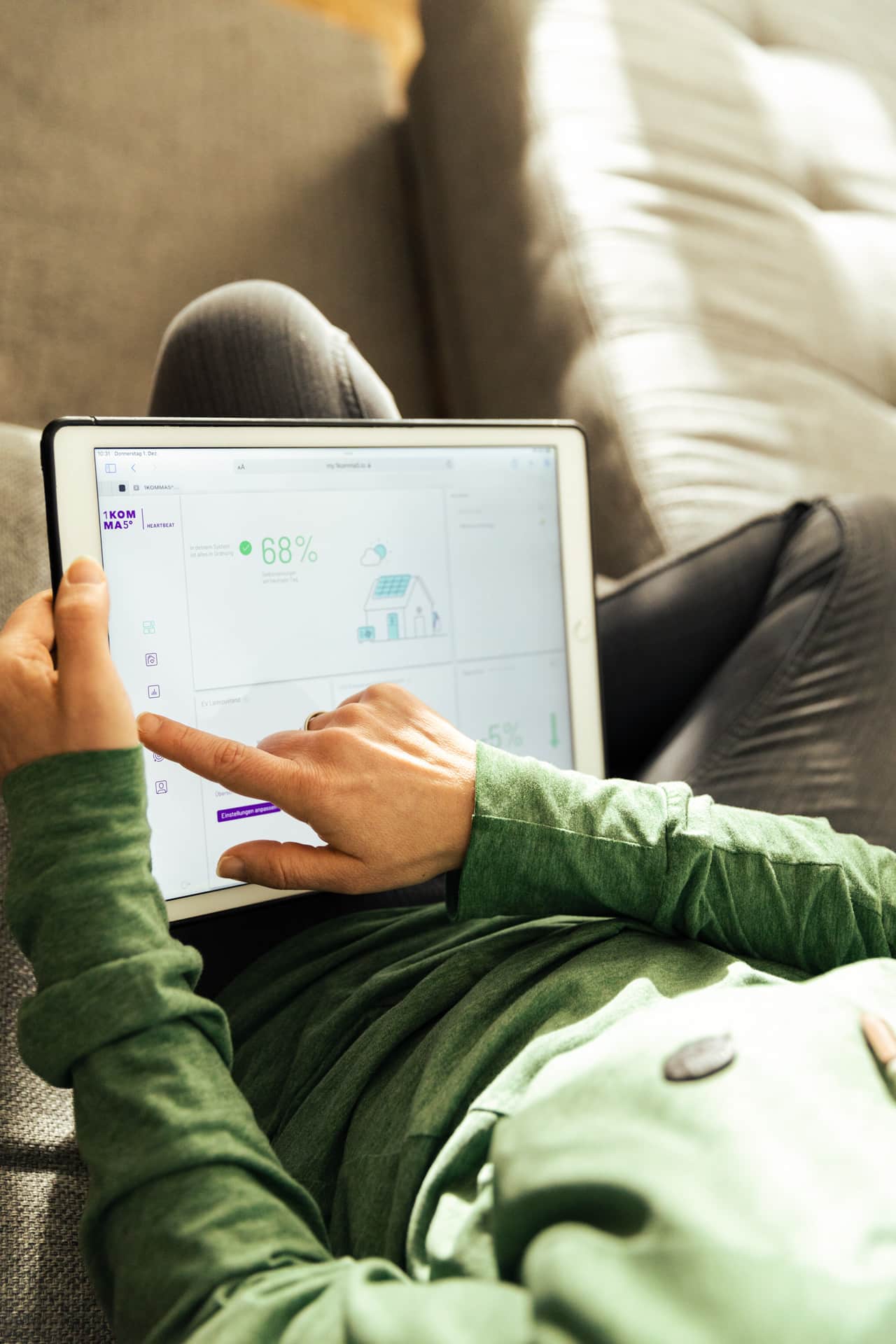 Nainen vihreässä paidassa käyttää tablettia, jonka näytöllä näkyy kodin energiankulutustietoja ja 68 %\:n tehokkuusindikaattori, istuen harmaalla sohva.
