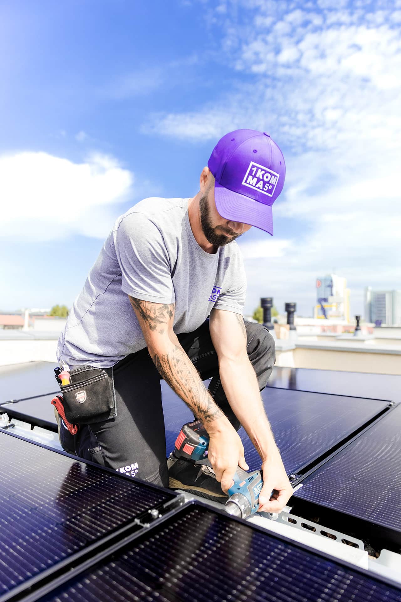 Kattoasentaja violetissa lippalakissa poraa kiinnittääkseen aurinkopaneelit sinisen taivaan alla.