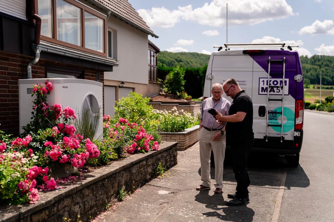 Kaksi miestä seisoo talon vieressä keskustellen valkoisen, violetilla tehosteväreillä varustetun pakettiauton lähellä lämpöpumpusta. Heidän vierellään kukkii kirkkaan pinkkejä kukkia aurinkoiselta taivaalta.