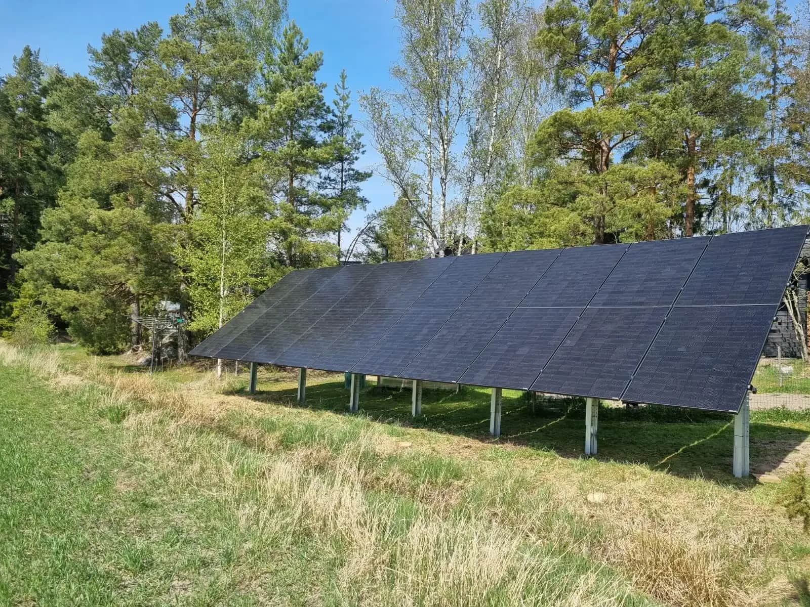 Maahan asennettu aurinkopaneelijärjestelmä metallisilla telineillä nurmialueella puiden vieressä sinisen taivaan alla.