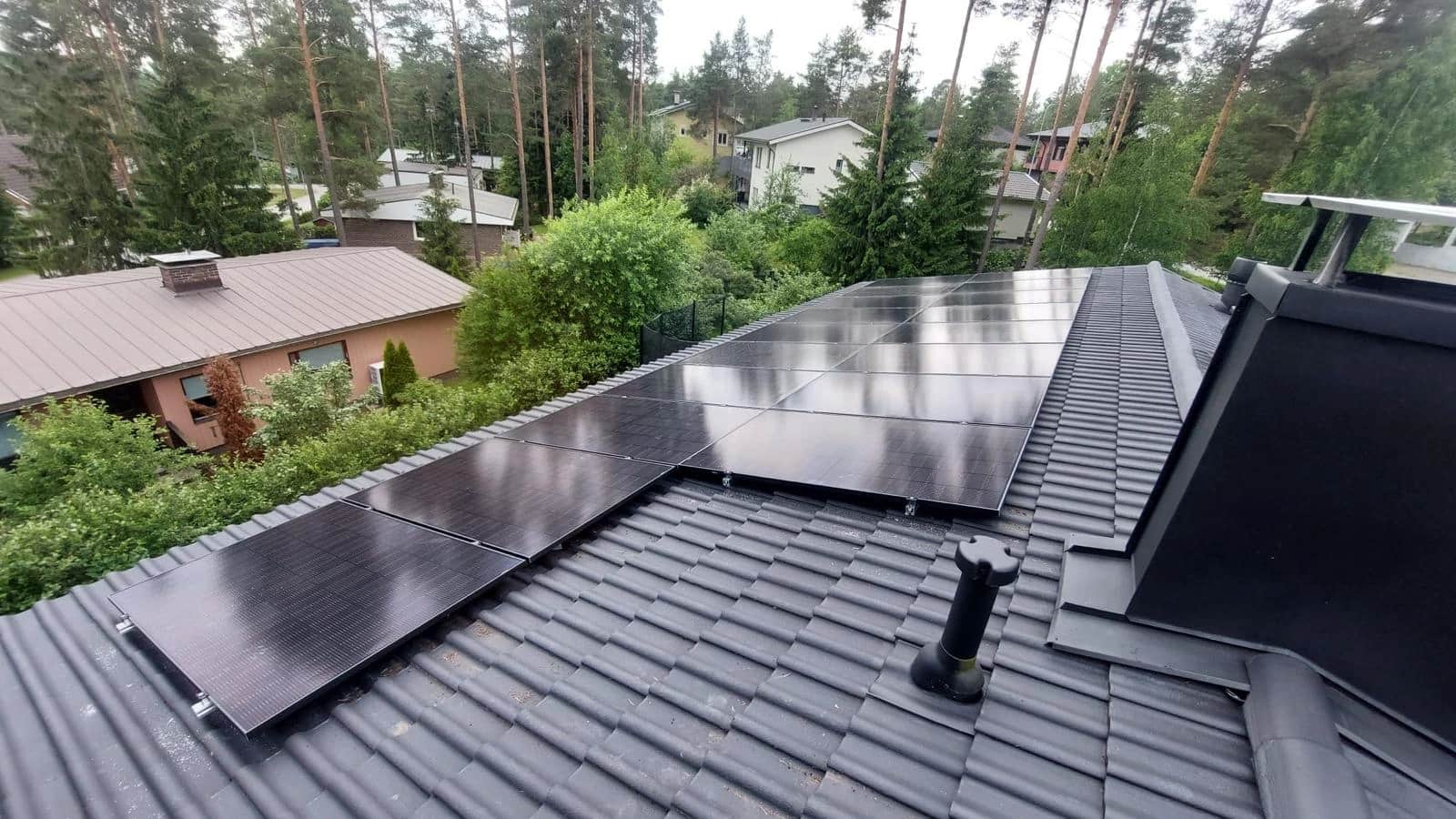 Aurinkopaneelit asennettuna tiilikatolle, puiden ja asuinrakennusten ympäröimänä pilvisen taivaan alla.