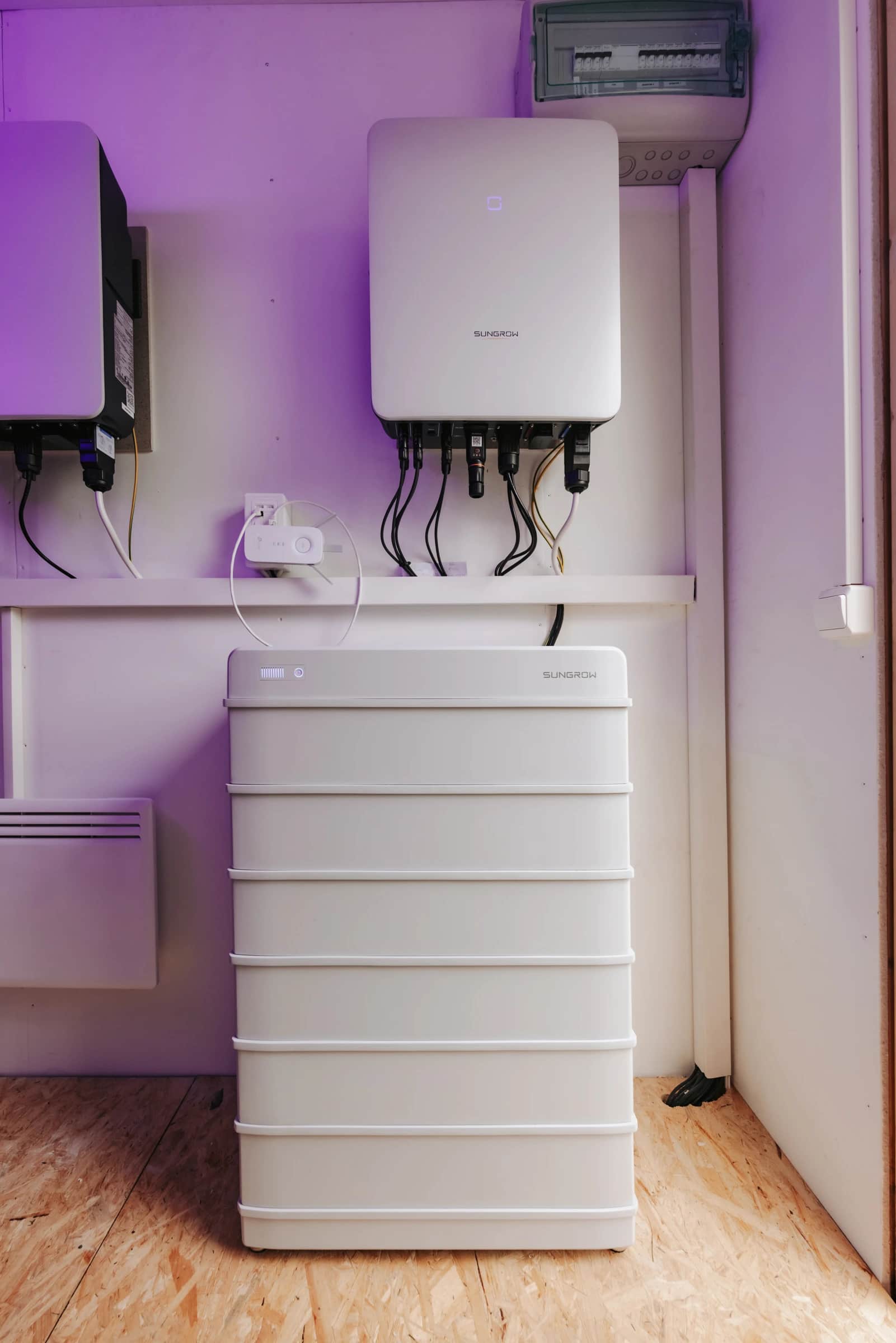 Valkoinen seinälle asennettu invertteri, joka on kytketty alapuolella olevaan suureen akkuvarastoyksikköön, huoneessa, jossa on puulattia.