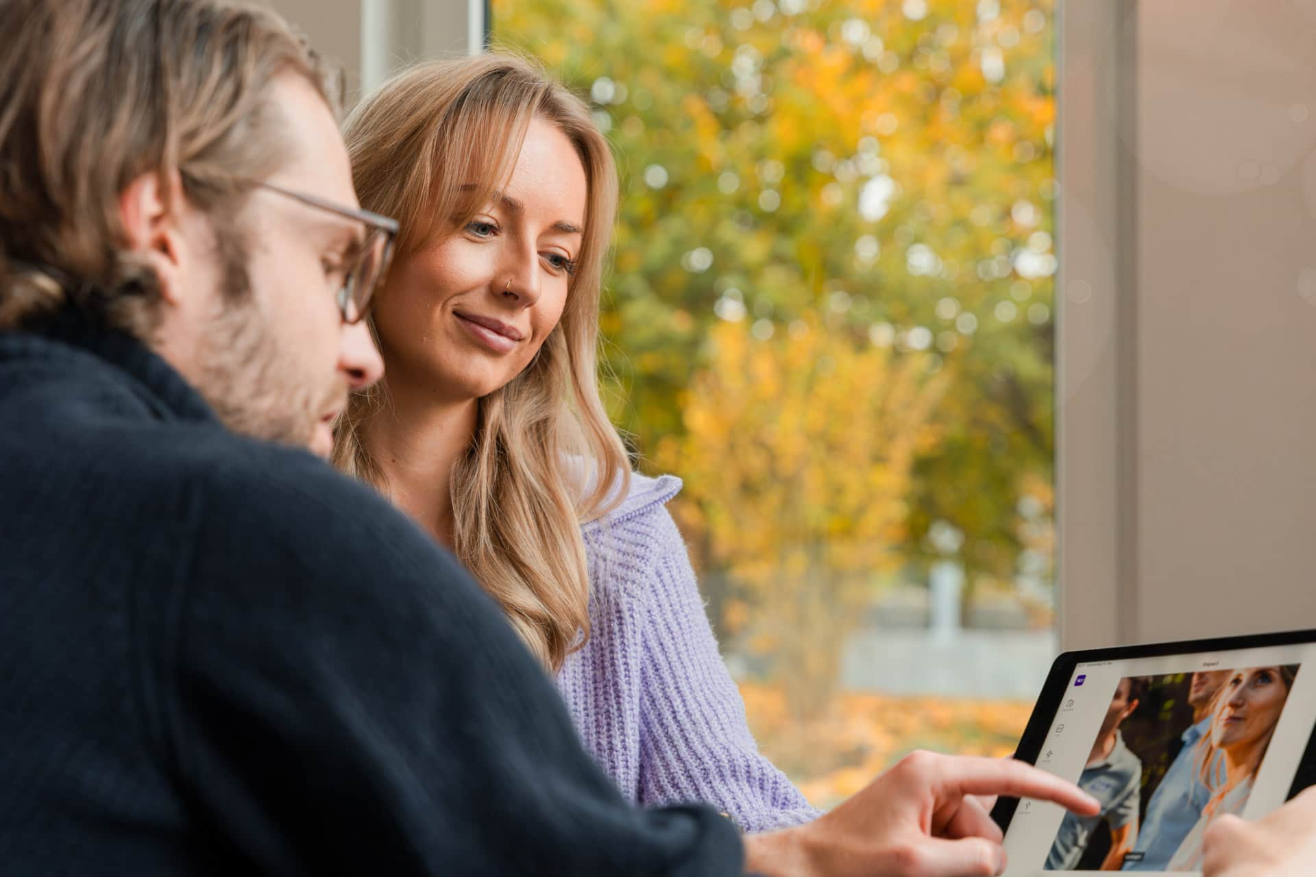 Mies ja hymyilevä nainen ikkunan äärellä katsovat valokuvia tabletista, mies osoittaa näyttöä; ulkona näkyy syksyisiä puita.