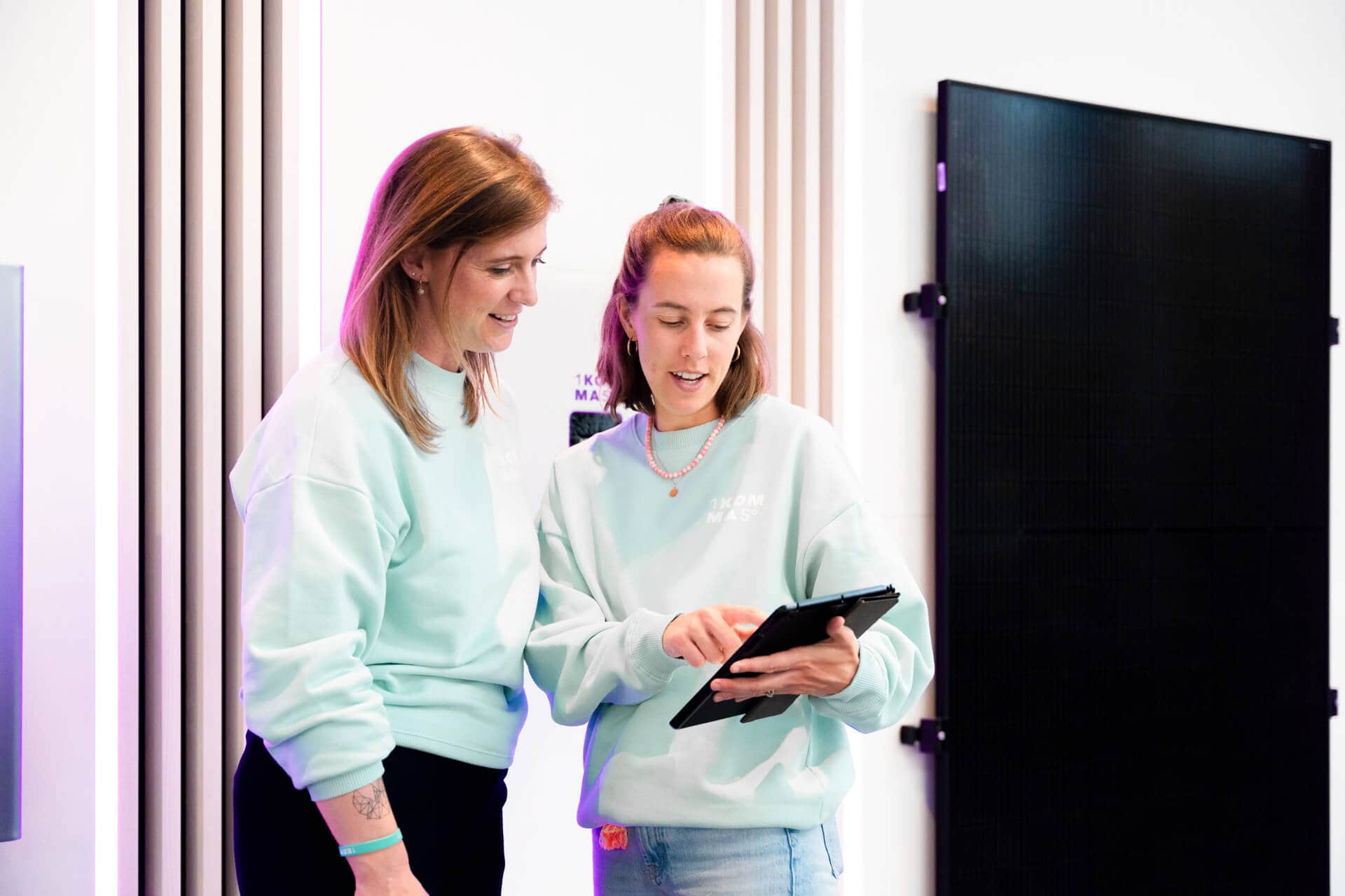 Kaksi rennosti pukeutunutta naista keskustelevat tabletin äärellä sisätiloissa; taustalla näkyy näyttö ja pystysuuntaisia aurinkopaneeleita.