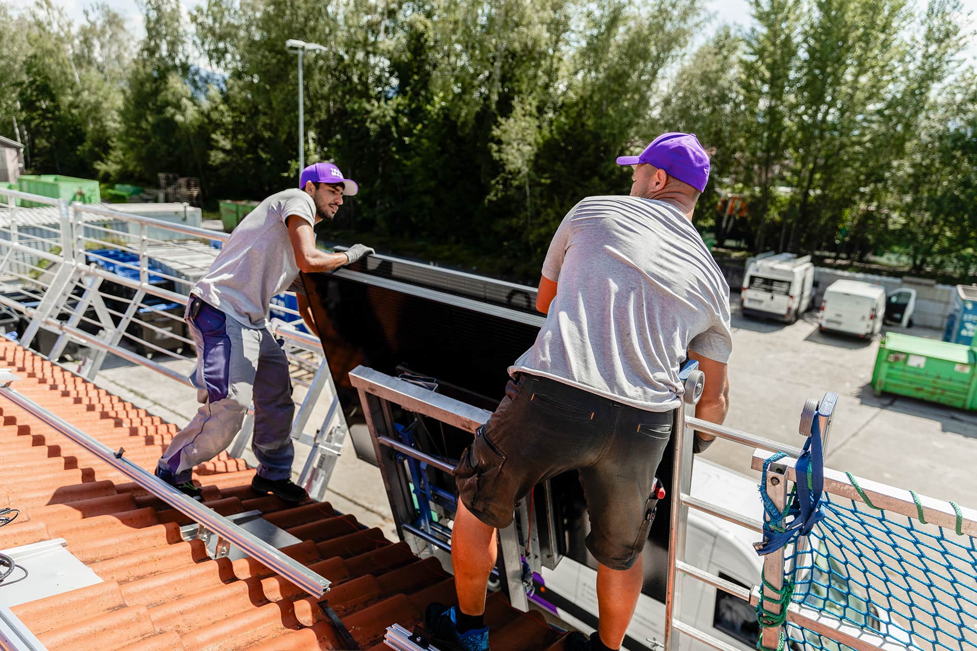 Kaksi työntekijää violetit lippikset päässään asentaa aurinkopaneelia kattolle. Taustalla näkyy puita ja pysäköityjä ajoneuvoja.