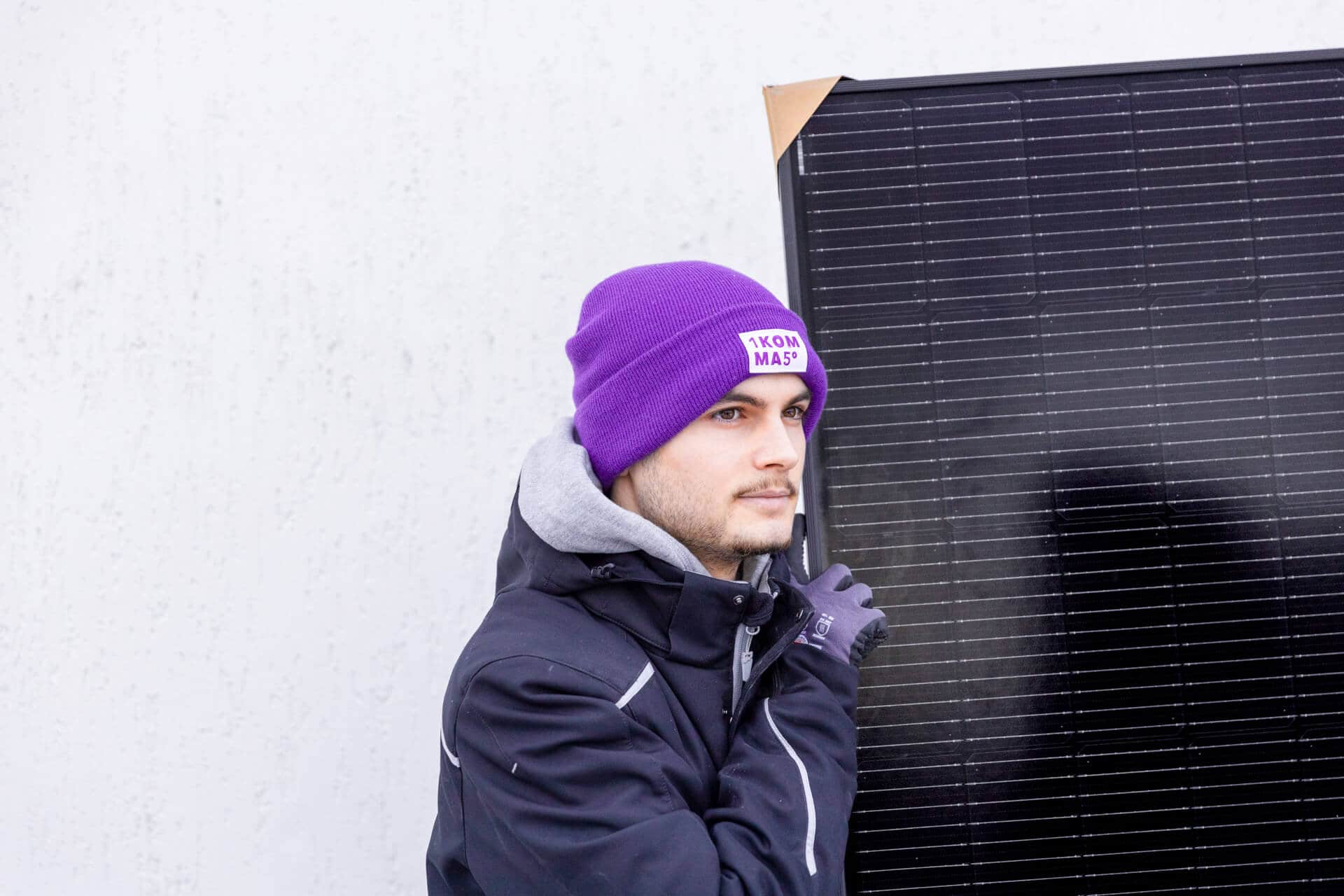 Henkilö violetissa pipossa ja tummassa takissa pitelee suurta aurinkopaneelia vaaleaa seinää vasten.