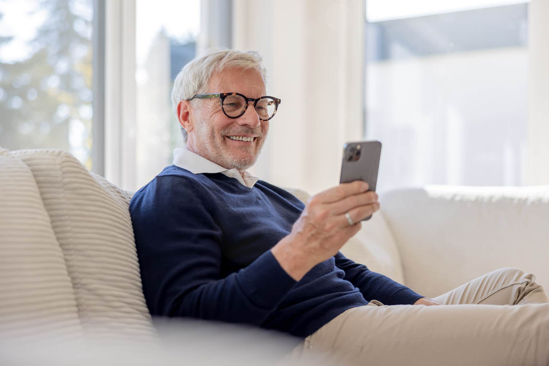 Iäkkäämpi mies silmälaseissa hymyilee älypuhelimelleen istuessaan sohvalla valoisassa huoneessa.