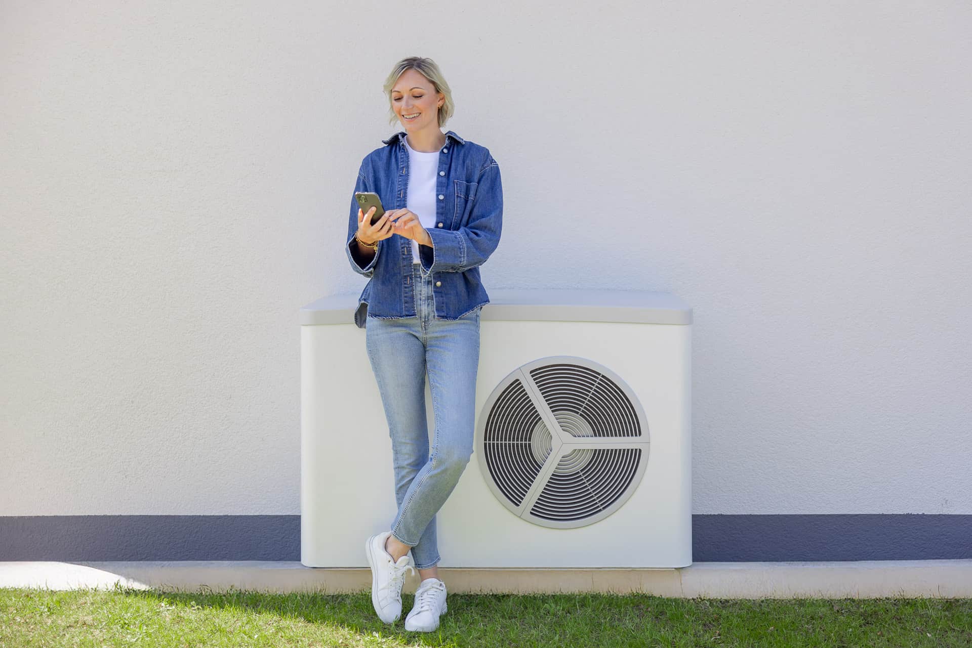 Nainen farkkuasussa nojaa ulkona olevaan ilmastointilaitteeseen ja katsoo puhelintaan, taustalla valkoinen seinä ja nurmikko.
