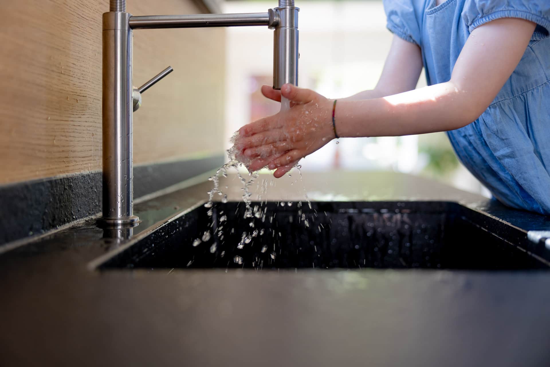 Lapsi pesee käsiään modernin hanan alla keittiön tiskialtaassa, veden roiskuessa.