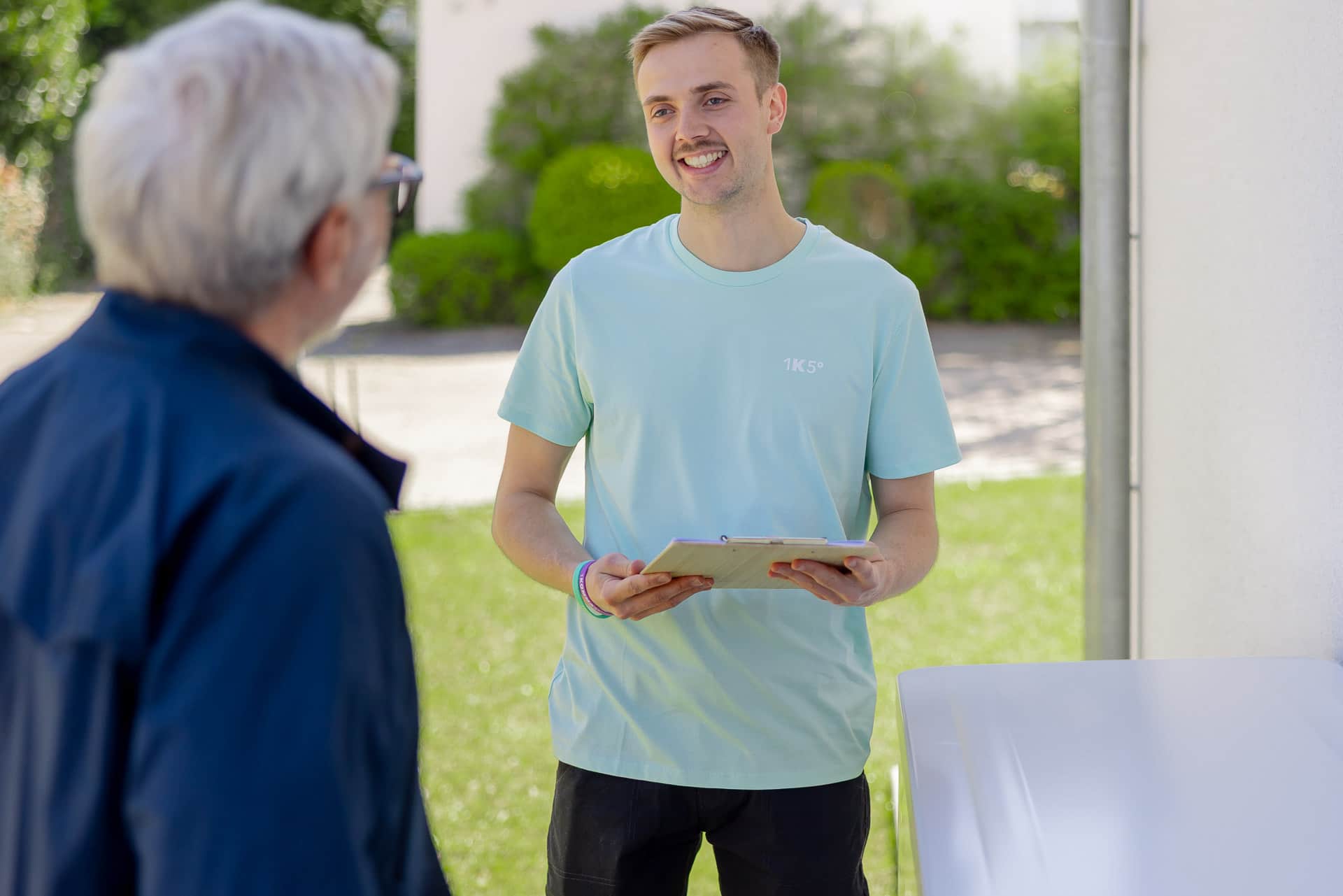 Hymyilevä mies vaaleansinisessä paidassa pitelee kansiota ja juttelee toisen henkilön kanssa ulkona aurinkoisena päivänä.