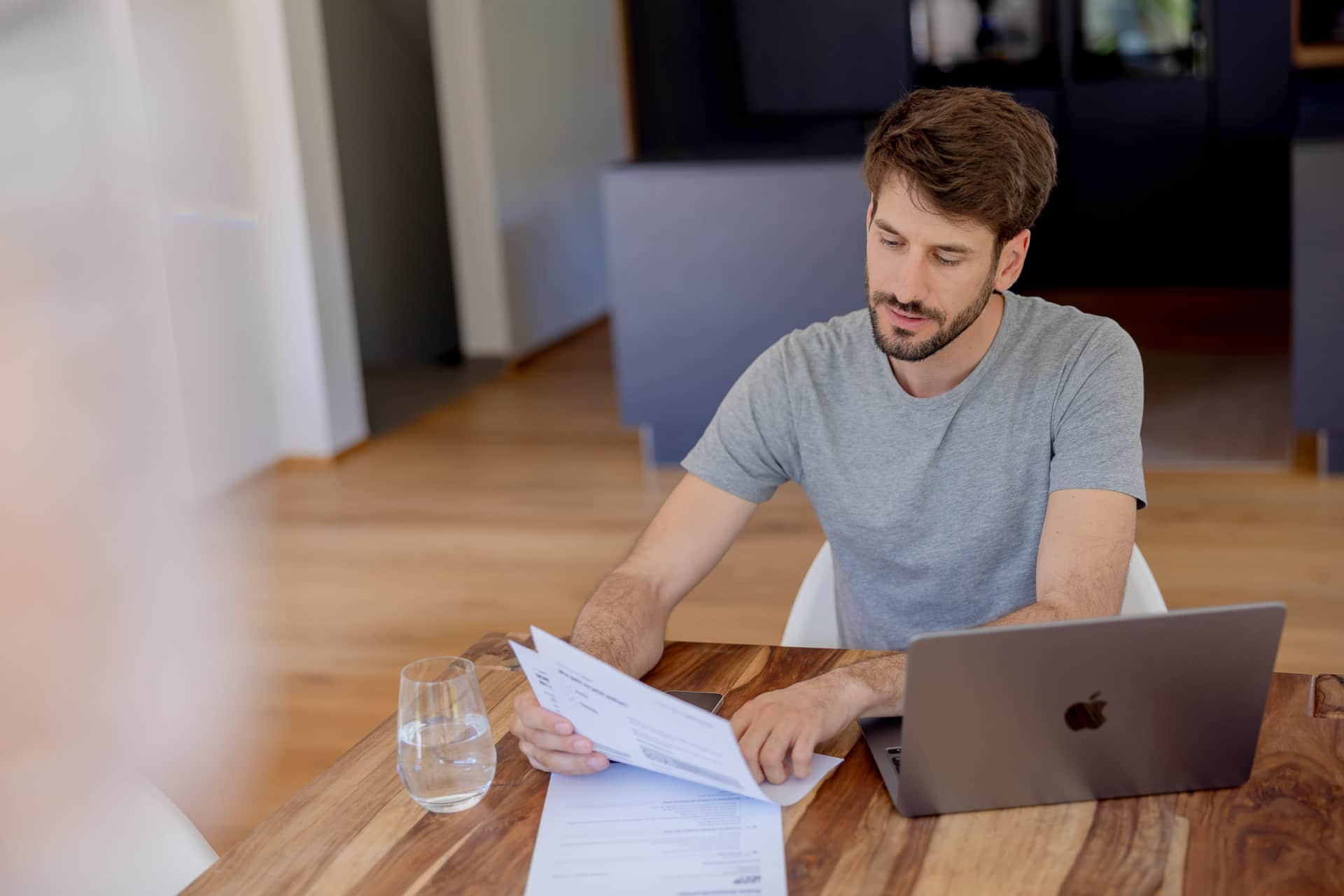 Mies istuu kotona puisella pöydällä tarkastelemassa asiakirjoja, edessään kannettava tietokone ja lasi vettä.