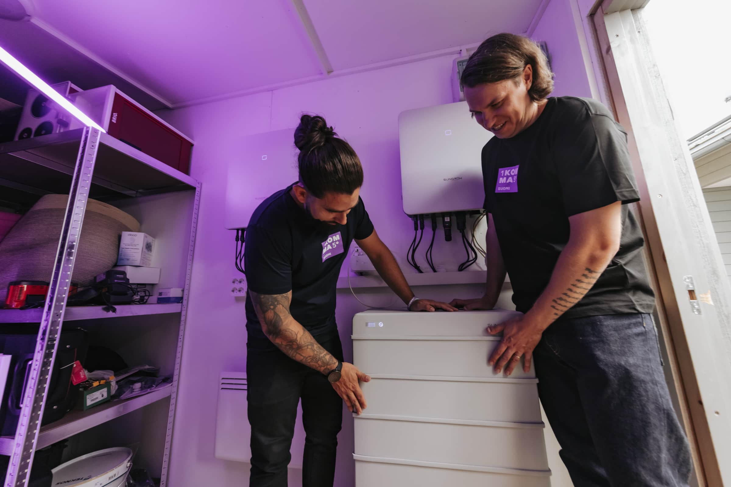 Kaksi ihmistä asentaa suuren akustojärjestelmän huoneeseen, jossa on hyllyjä. Valaistus on violetinsävyinen.