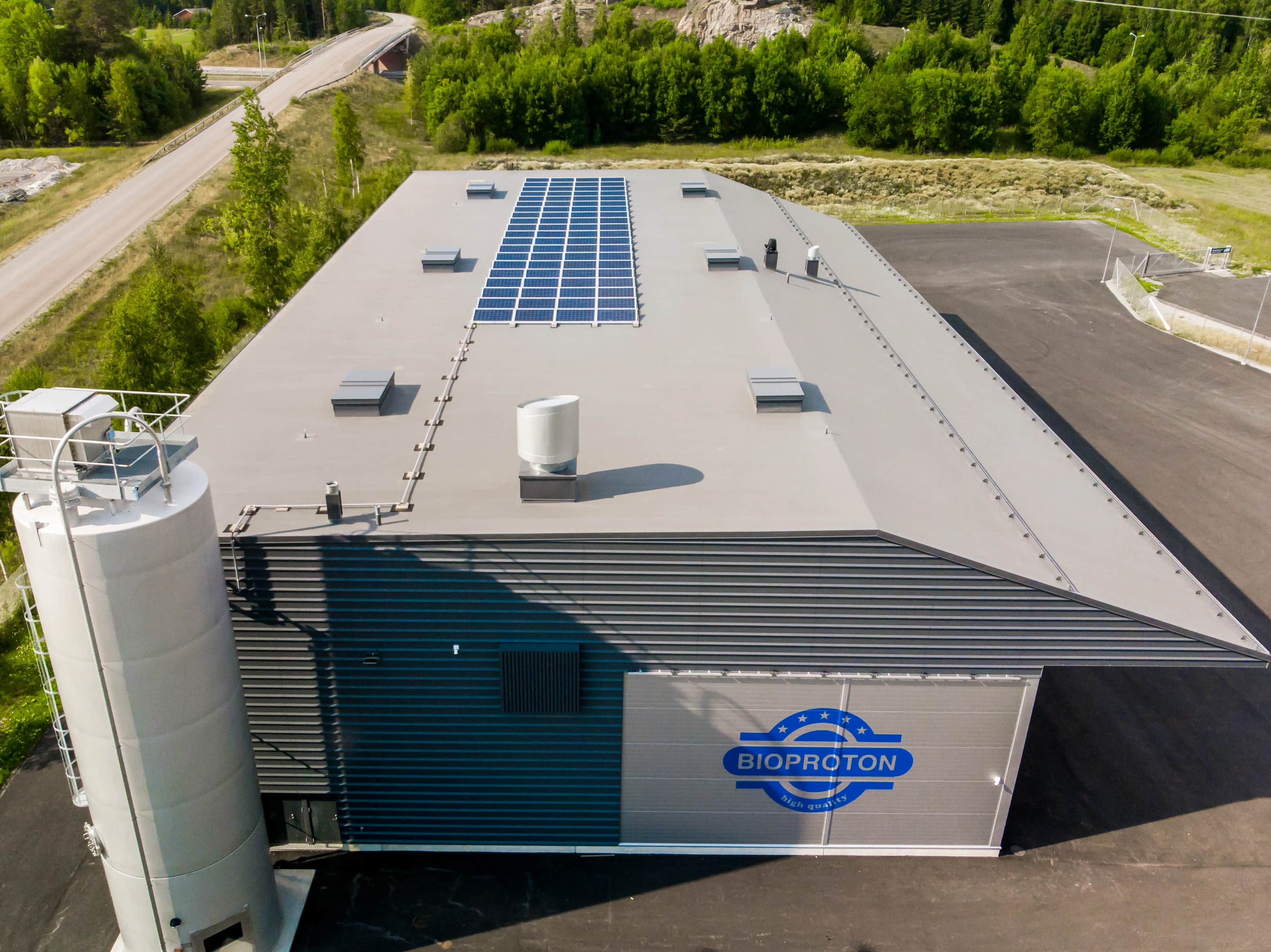 Ilmakuva harmaasta teollisuusrakennuksesta, jonka katolla on aurinkopaneelit, vieressä valkoinen siilo sekä ympärillä puita ja kulkuteitä.