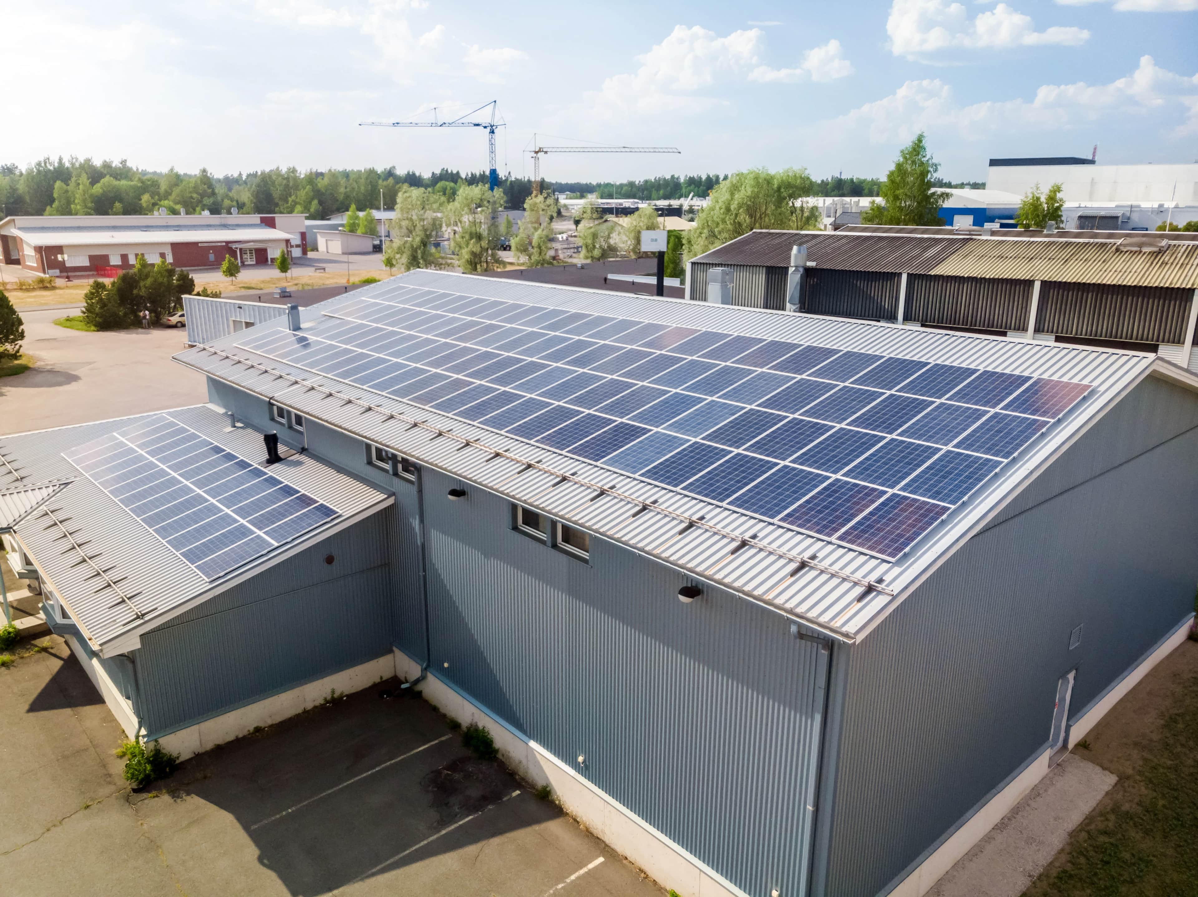 Ilmakuva teollisuusrakennuksesta, jonka metallikatoilla on laajat aurinkopaneelijärjestelmät, ympärillä varastoja ja puita sinisen taivaan alla.
