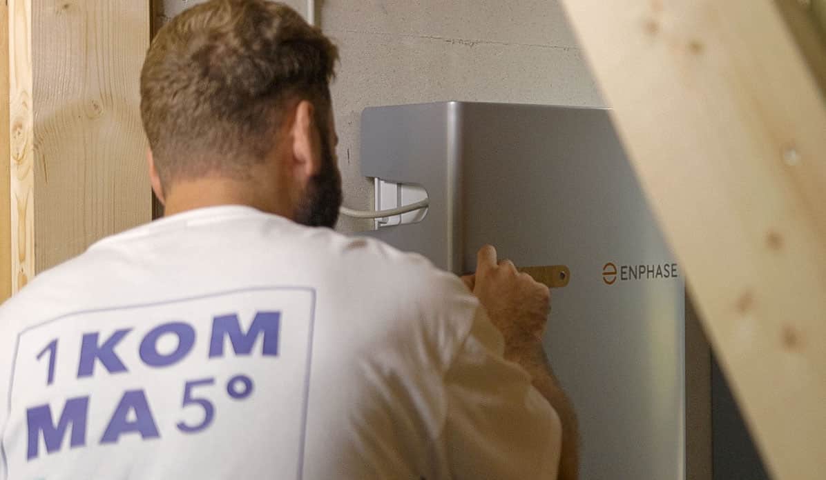 Henkilö valkoisessa 1KOMMA5°-paidassa asentaa Enphase-aurinkoenergian varastointilaitetta seinälle työkalua käyttäen, osittain puurungolla varustetussa huoneessa.
